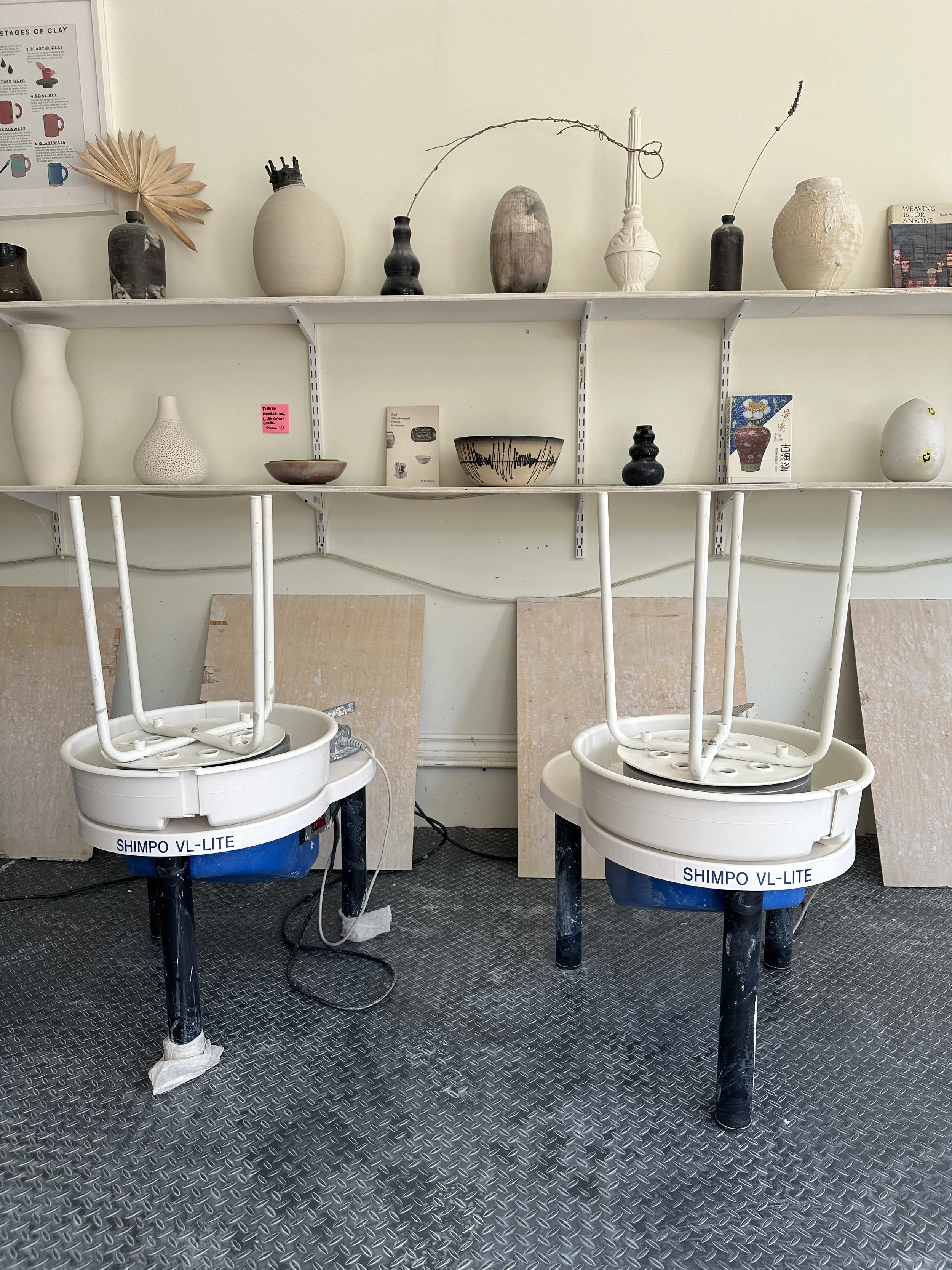 Two pottery wheels with white bases and black legs in a pottery studio, with shelves of various ceramic vases and bowls behind them, and a textured metal floor.