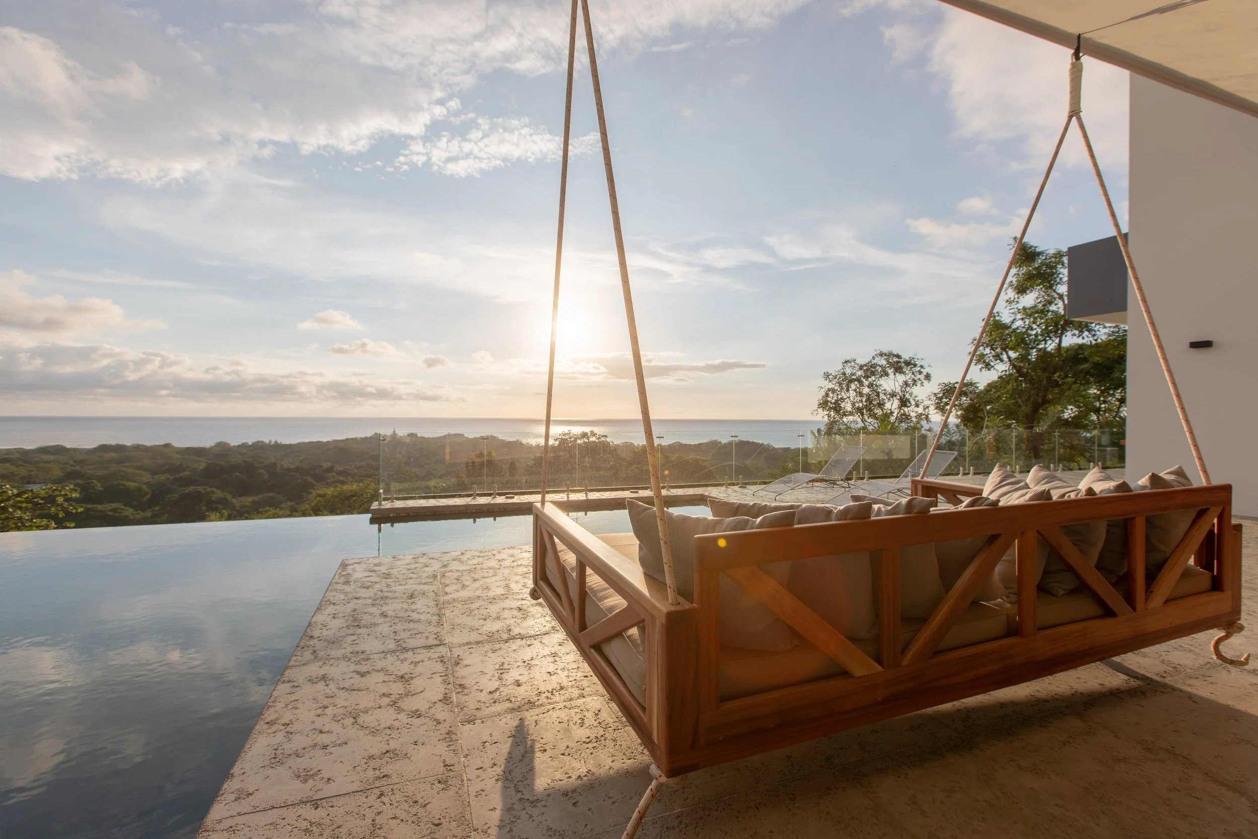 Outdoor lounge area with a wooden swing sofa overlooking an infinity pool and scenic landscape at sunset.