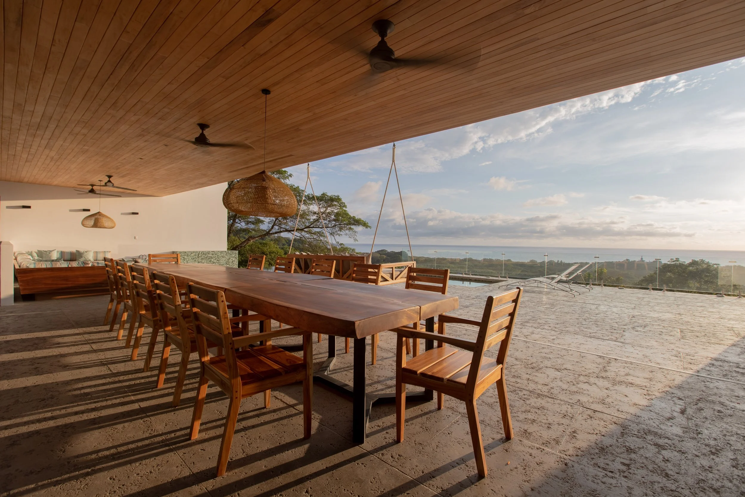 Outdoor dining area with wooden table and chairs, ceiling fans, and ocean view.