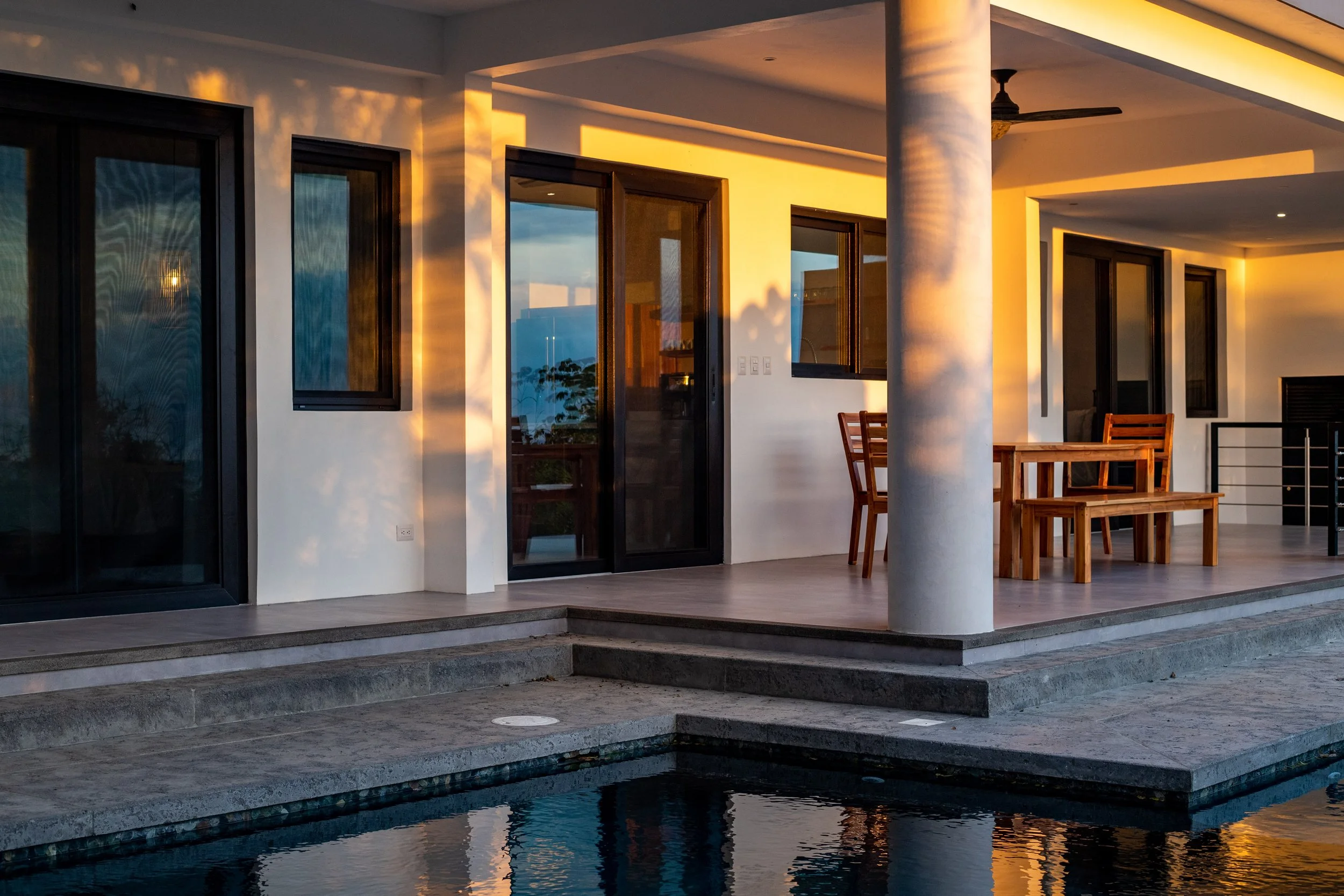 Modern patio with wooden furniture and poolside view, highlighted by warm evening sunlight.