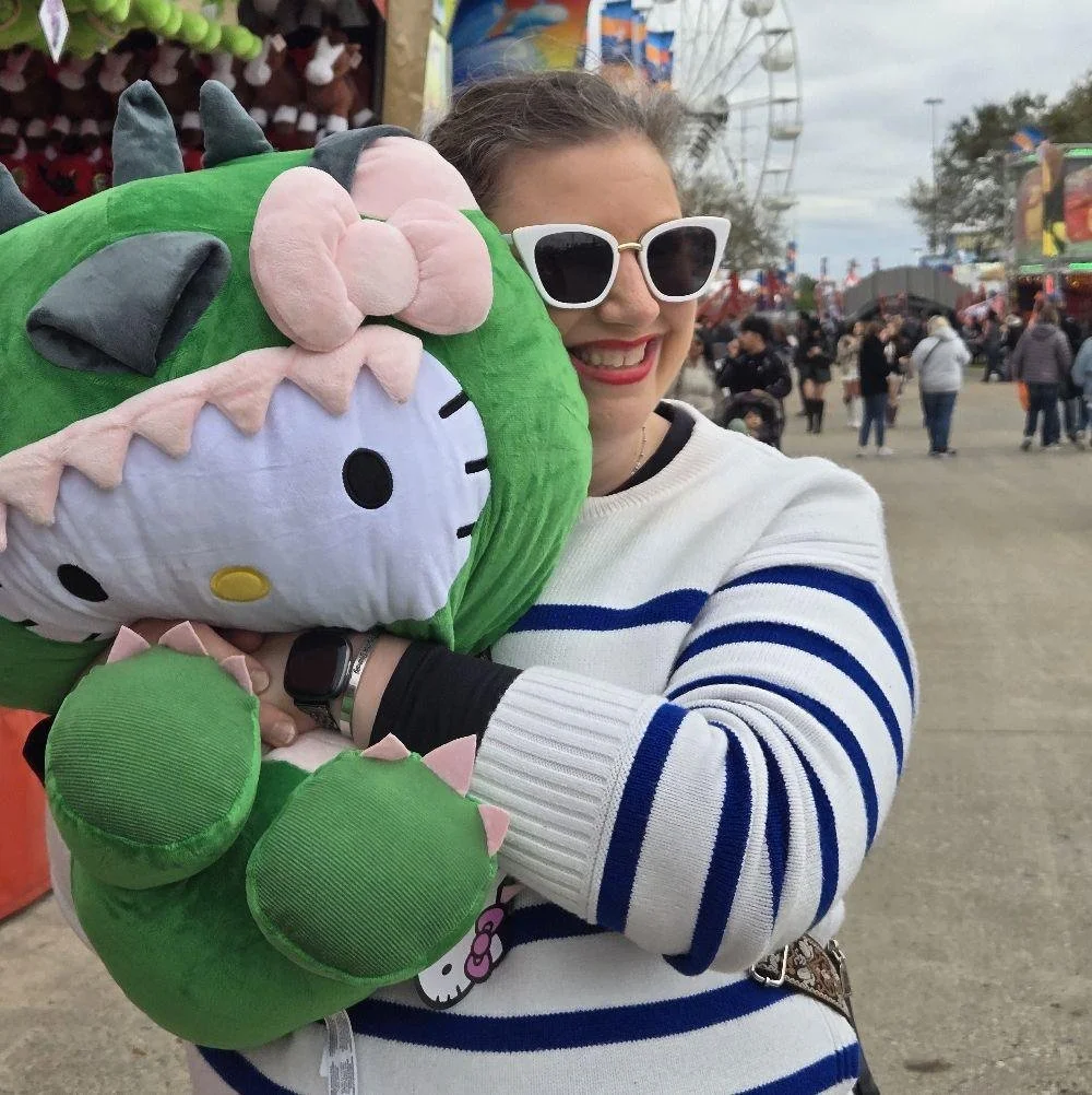 Person with sunglasses hugging a large green dinosaur-themed plush toy, with a carnival scene in the background.