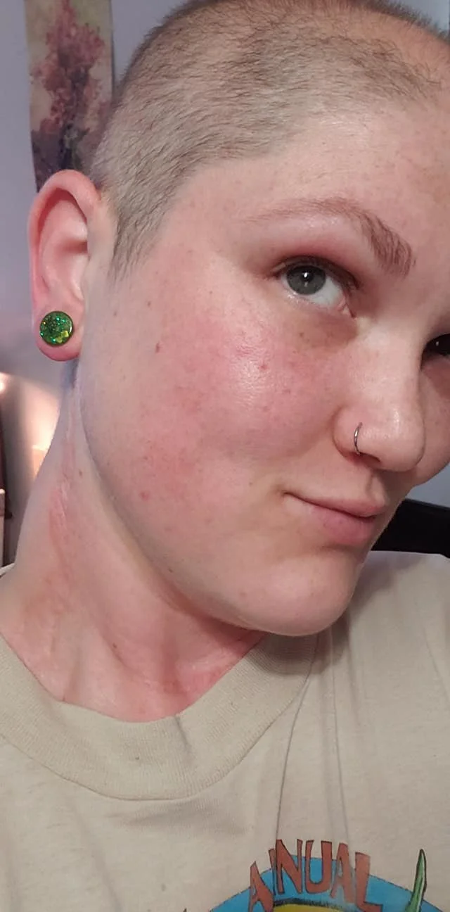 Person with shaved head, wearing a green earring and a nose ring, looking to the side.