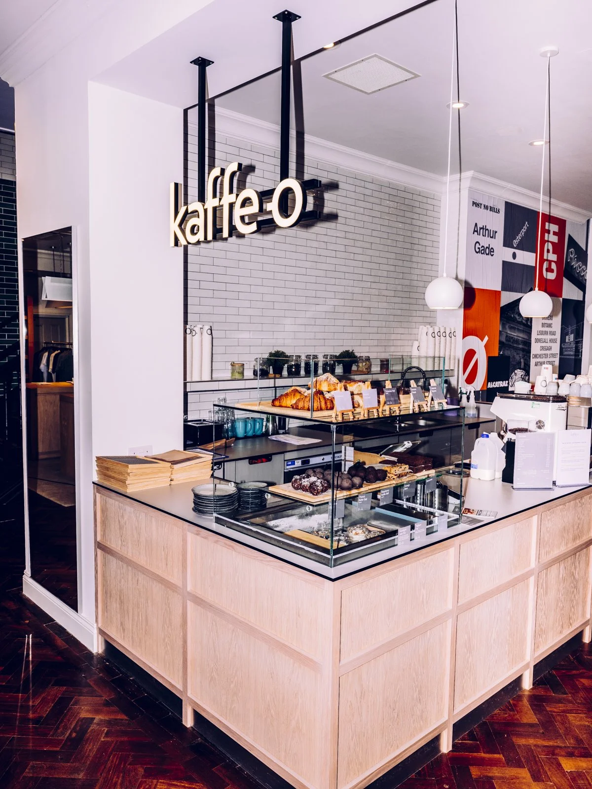 Coffee shop counter with pastries and baked goods, including croissants and chocolate balls, on display. The sign says 'kaffe-o' and there are hanging white pendant lights. Background has a white brick wall and decorative banners.
