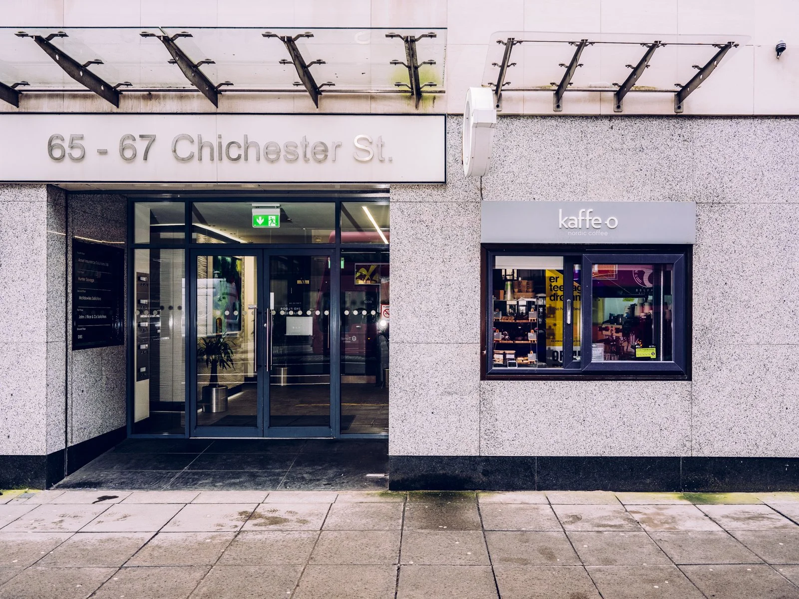 A storefront with a sign indicating 65-67 Chichester Street. The entrance has glass sliding doors with a green exit sign above. Next to the entrance is a window displaying products and a sign reading 'kaffe o' for Nordic coffee.