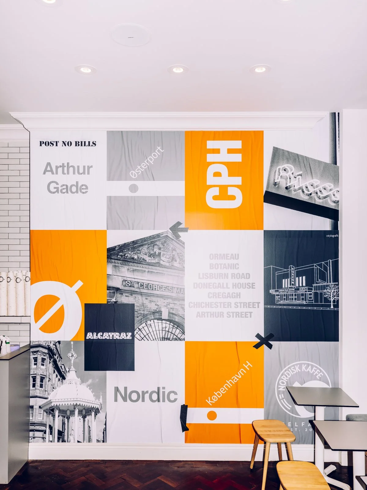 A colorful wall mural with a collage of images and text in gray, orange, white, and black. The mural features architectural elements, text such as "POST NO BILLS," "Arthur Gade," "NORDIC," and others, along with abstract graphics and a neon sign in t