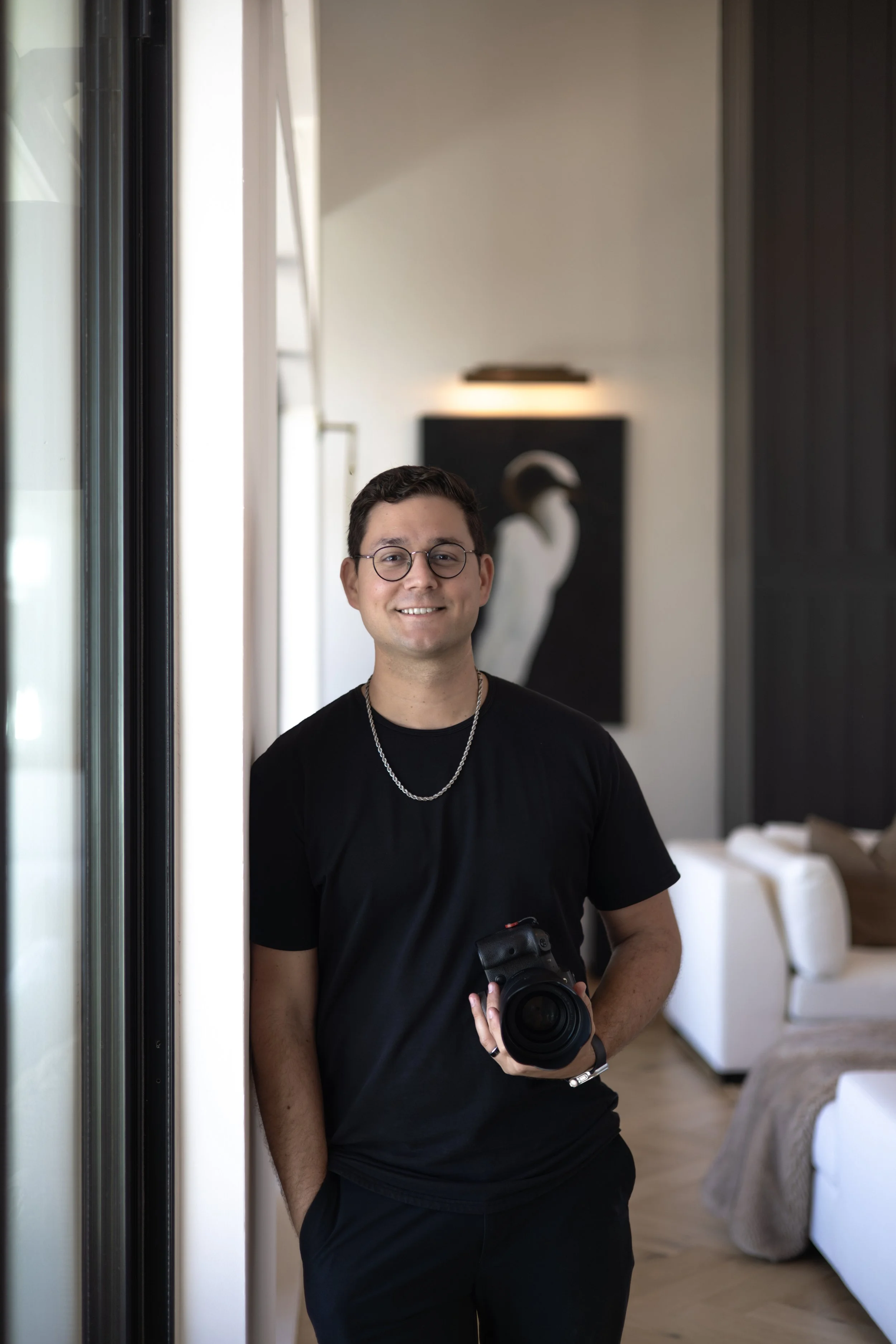 A young man with glasses and a silver chain necklace, holding a camera, standing indoors near a glass wall with a white couch and framed art in the background.
