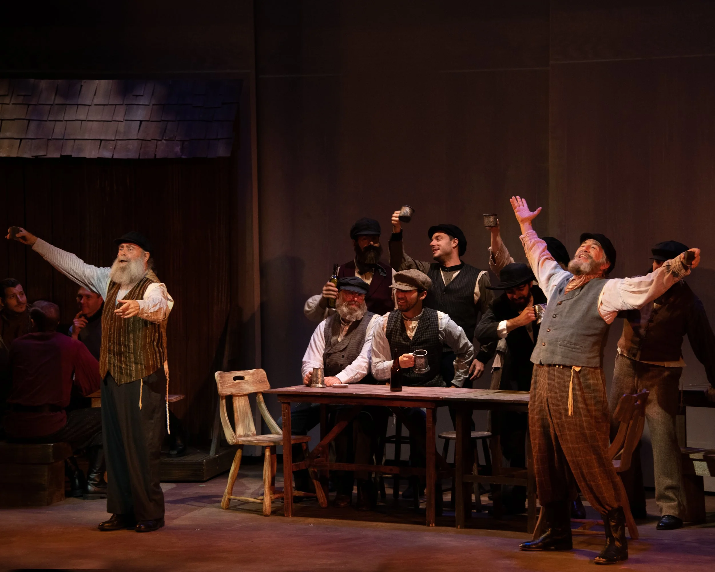 Theatrical performance with men in period costumes, possibly set in a bar or tavern. Two men stand at the front with their arms outstretched, while others are seated at a wooden table holding mugs. The scene conveys a lively, festive atmosphere.