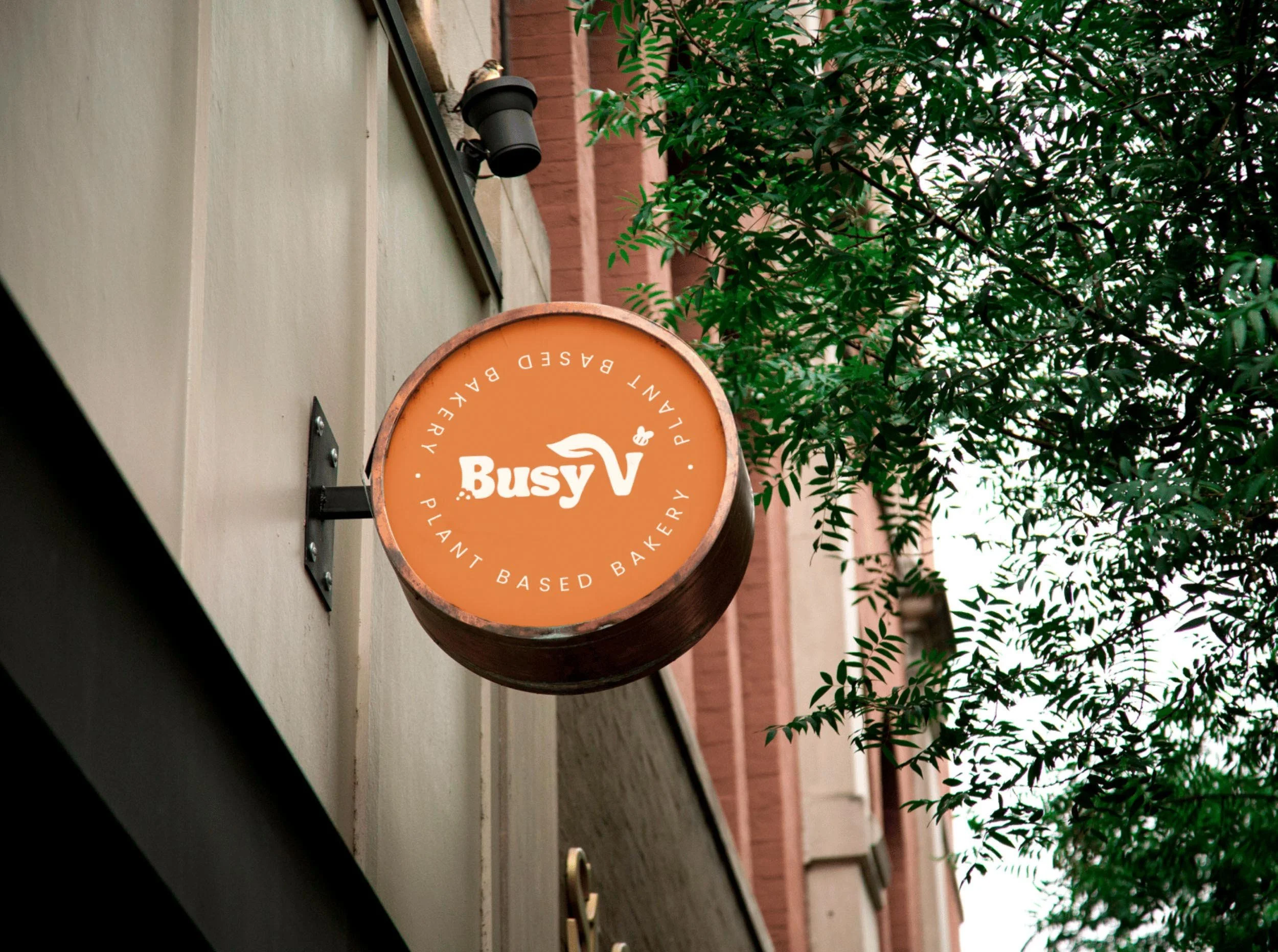 A mock-up of a round street sign for Busy V Bakery. The sign is orange and readys "Busy V" with smaller text "plant-based bakery" around the edges. The sign was designed by Sprout Design.
