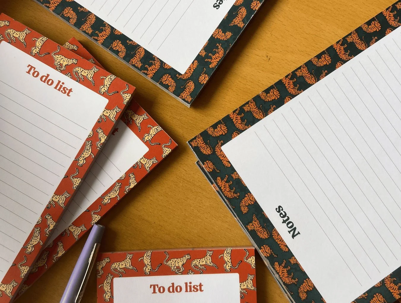 Notebooks and notepads with illustrated animal-themed borders, some labeled "To do list" and "Notes," on a wooden table with a purple pen.  The notepads were designed by Sprout Design.