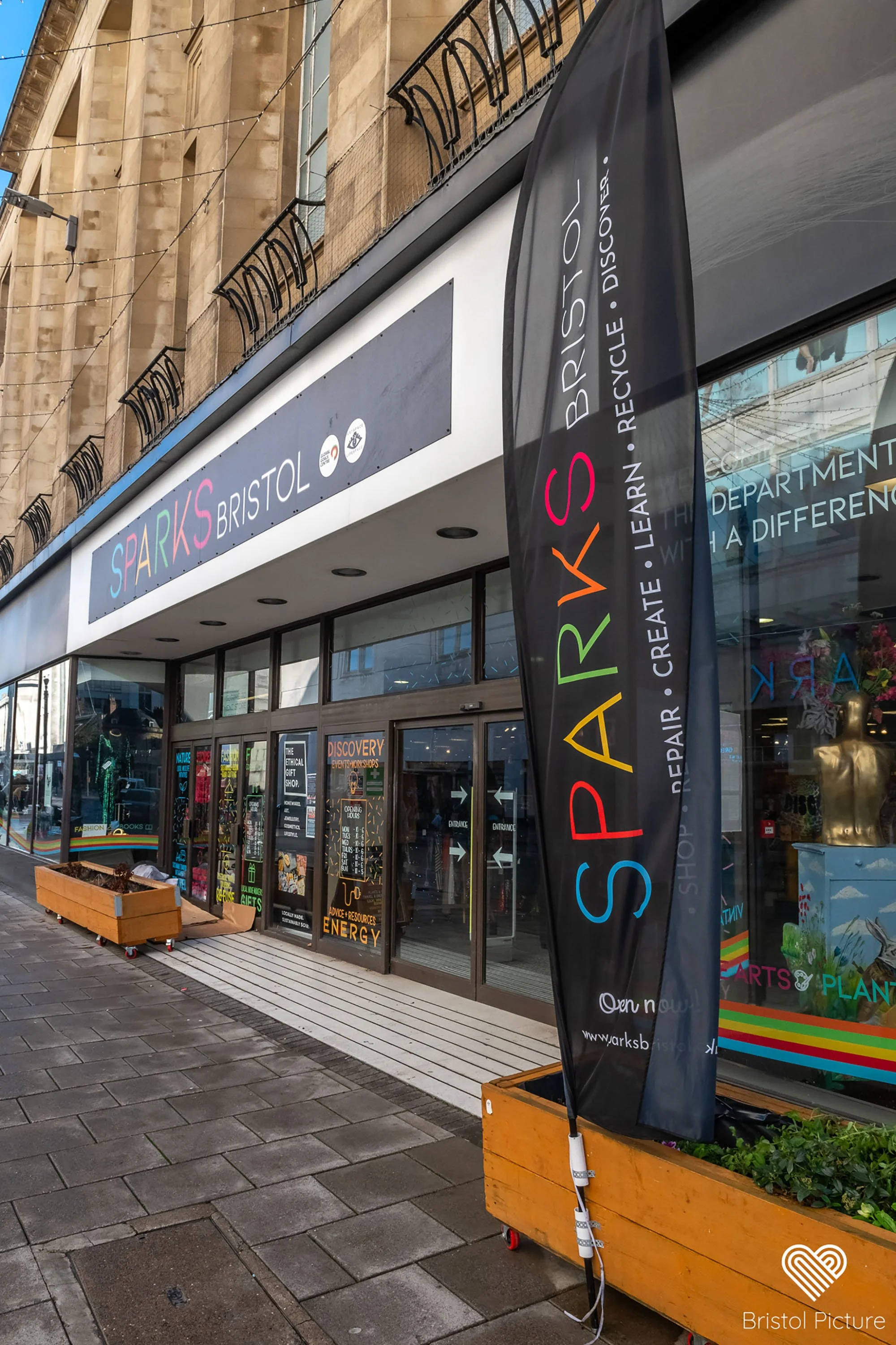 A storefront with signs for Sparks Bristol and a colorful banner advertising recycle, repair, learn, create, and discover activities. There's a wooden planter box with plants and a black flag with the same text. 