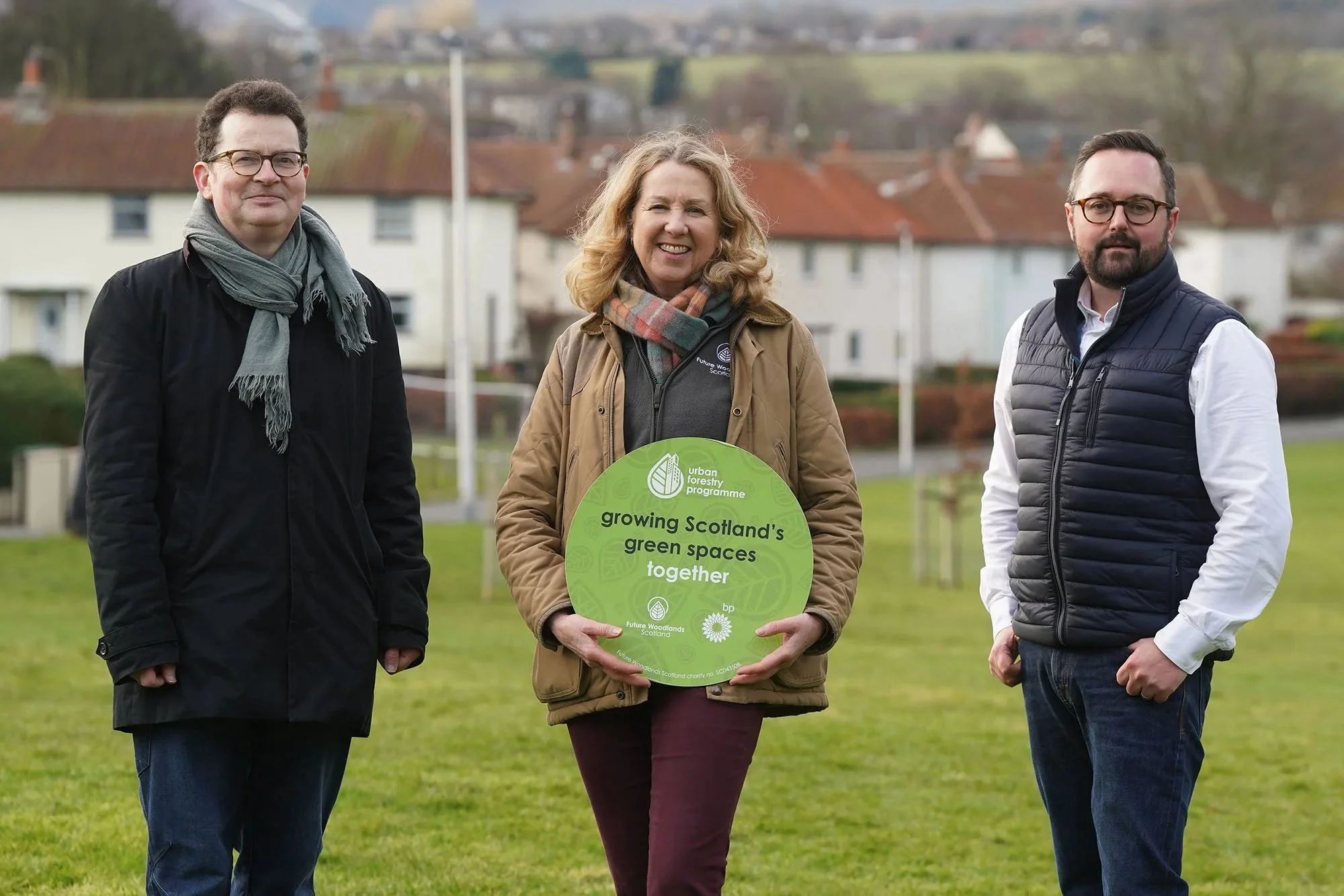 Three people standing outdoors on a grassy area during daytime, holding a green circular sign promoting the Urban Forestry Programme. The circular sign was designed by Sprout Design.