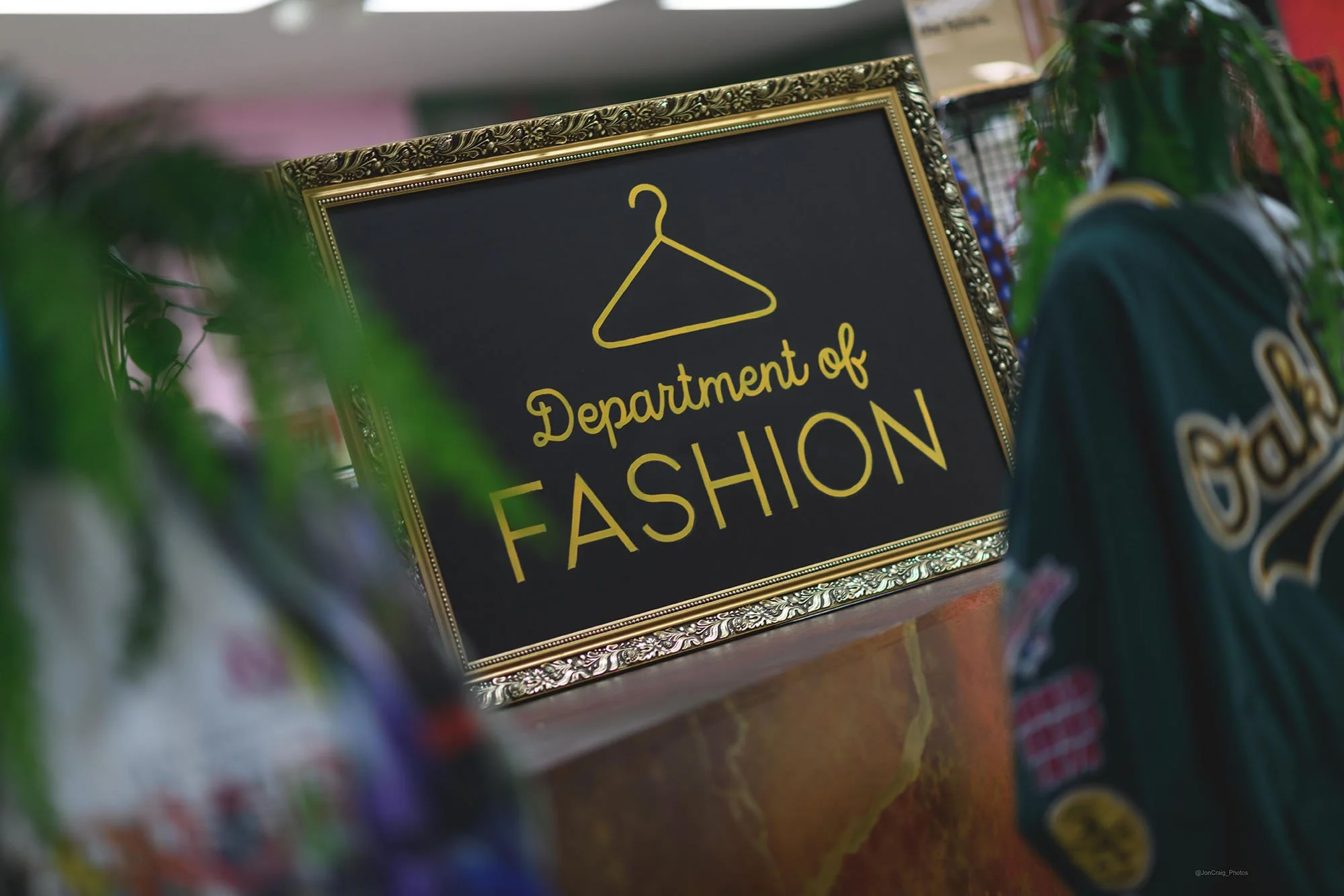 Decorative sign reading 'Department of Fashion' with a yellow hanger icon, framed in an ornate gold border, surrounded by blurred plants and clothing items inside of Sparks Bristol.