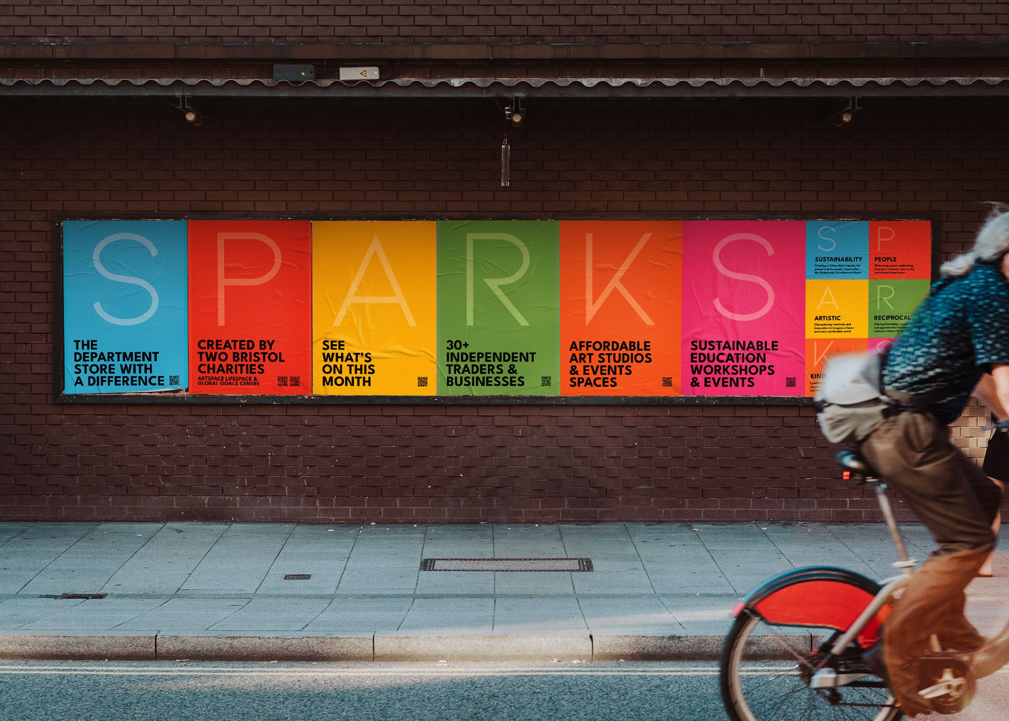 Colorful posters promoting Sparks Bristol on a brick wall with pedestrians walking and a cyclist passing by.