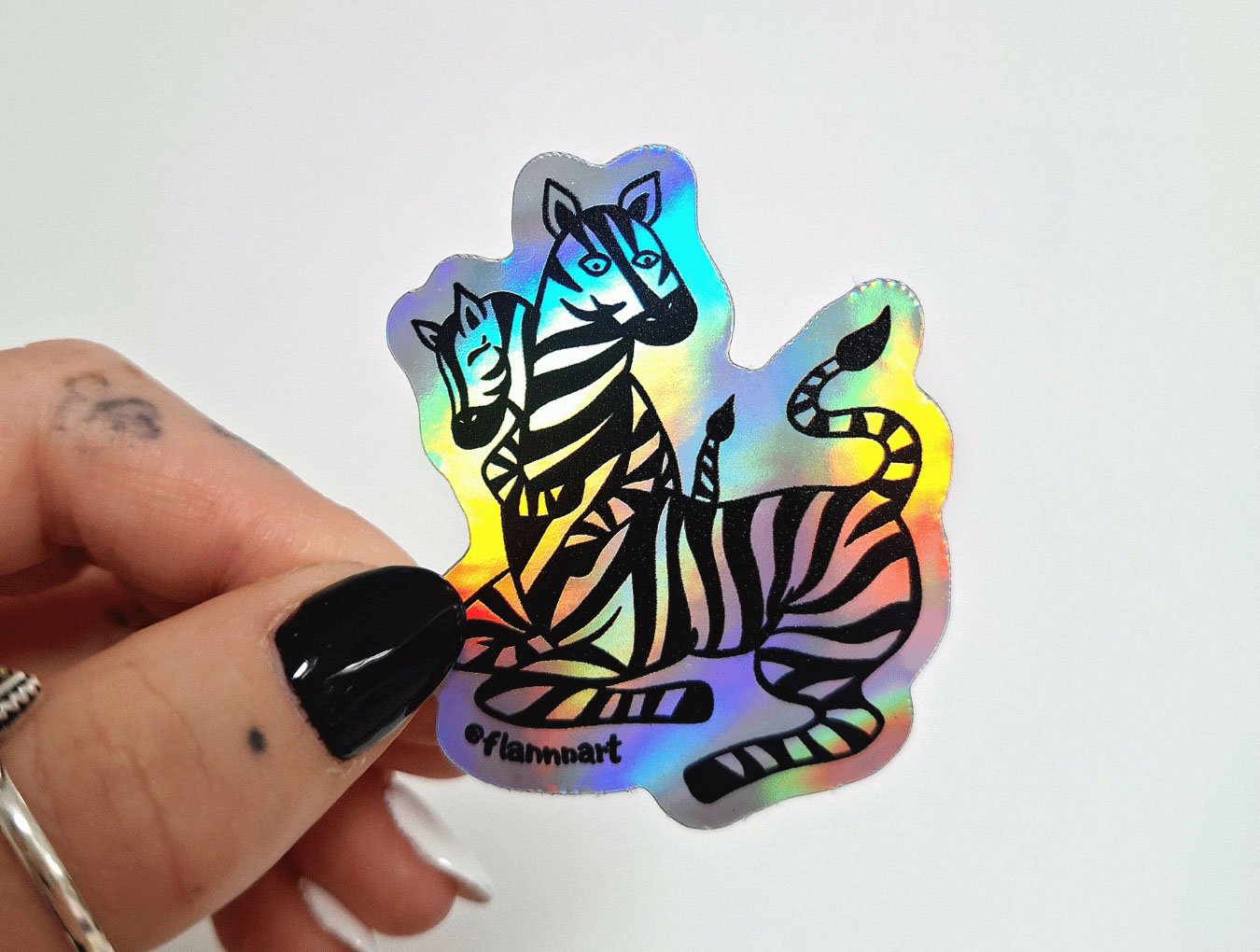 Holographic sticker of two black-outlined zebras, one adult and one foal, with the adult zebra sitting and the foal leaning on it. The sticker was designed by Sprout Design.
