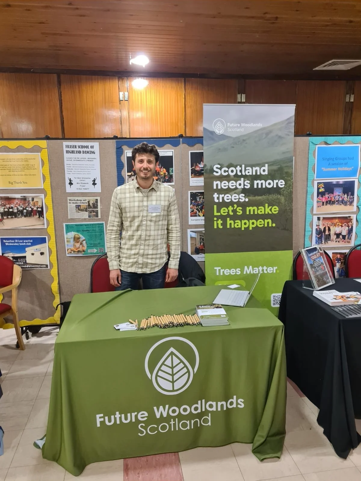 A man standing behind a table at a booth promoting Future Woodlands Scotland, with a large green banner that reads 'Scotland needs more trees. Let’s make it happen. Trees Matter.' The booth has pamphlets, pens, and a notebook on the table. 
