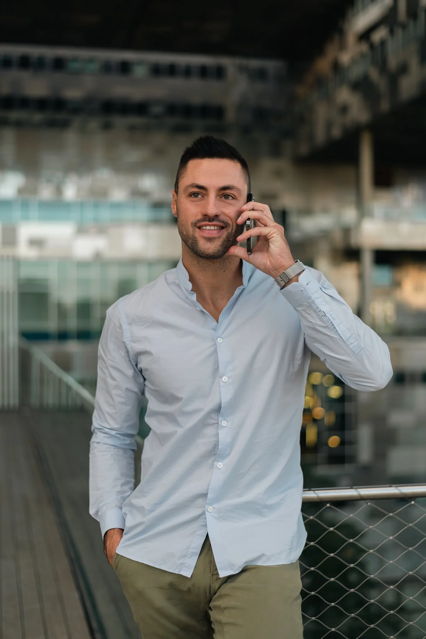 Man in light blue shirt and khaki pants talking on cellphone outdoors near modern building.