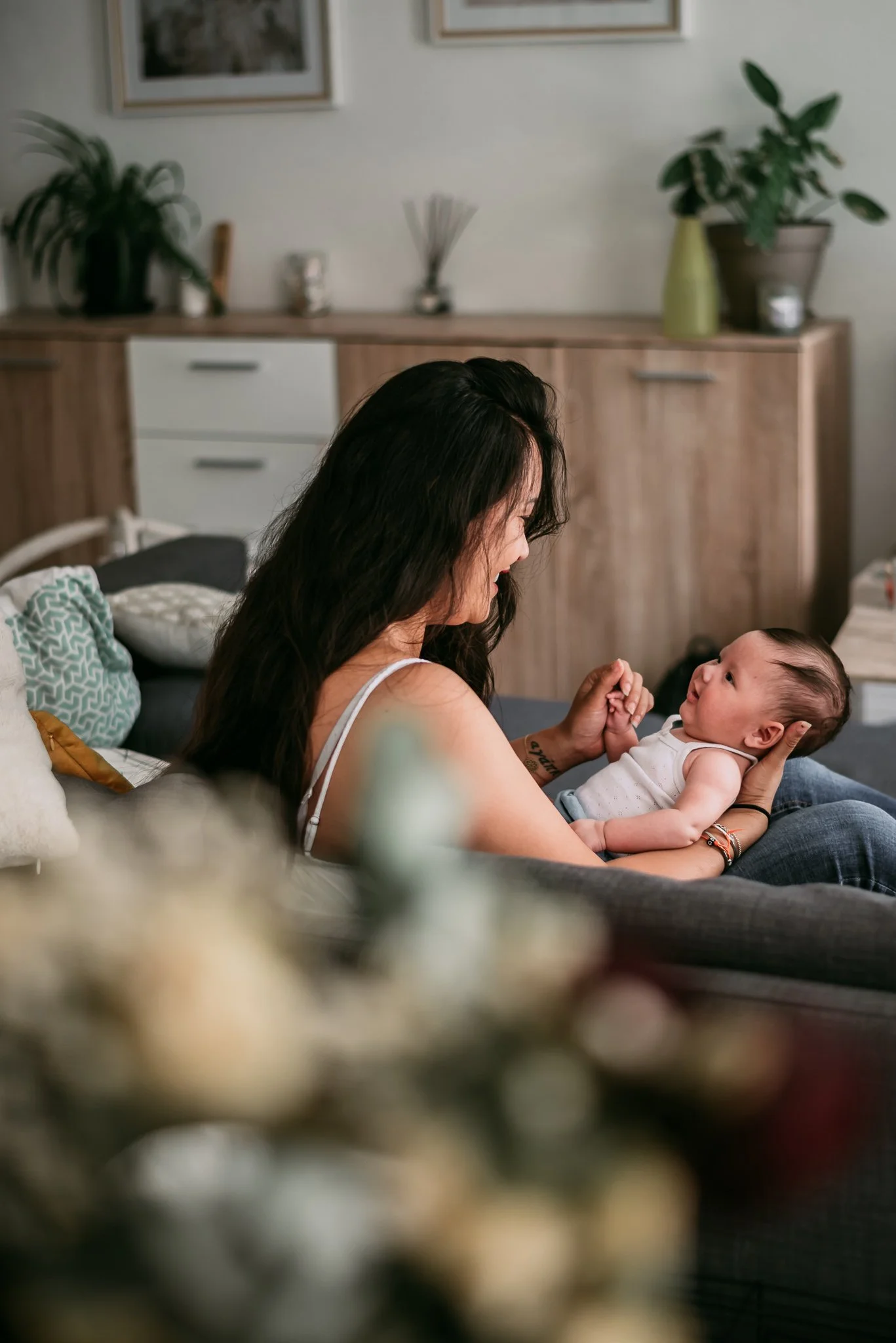 A woman with long dark hair holding a baby in a living room, both face to face, with the woman smiling at the baby. The background shows houseplants, framed pictures, and a wooden sideboard.