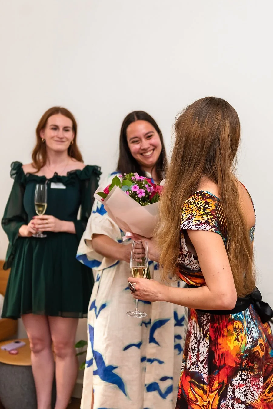 Three women at a social gathering, with one woman giving a bouquet of flowers and a glass of champagne to another woman, while the third woman stands in the background holding a glass of champagne and smiling.