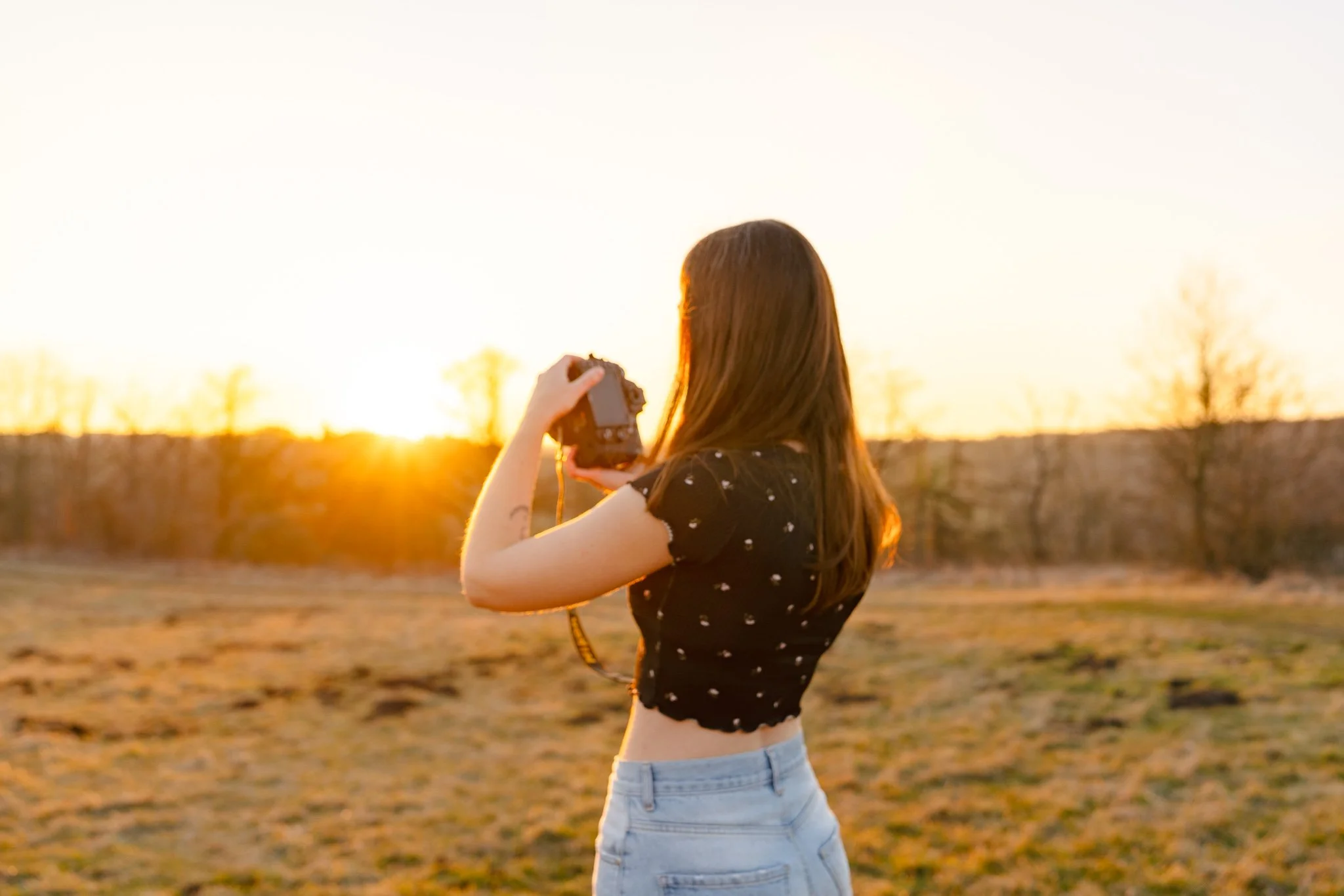 A woman with long brown hair wearing a black crop top and light blue jeans takes a photo with a camera outdoors during sunset.