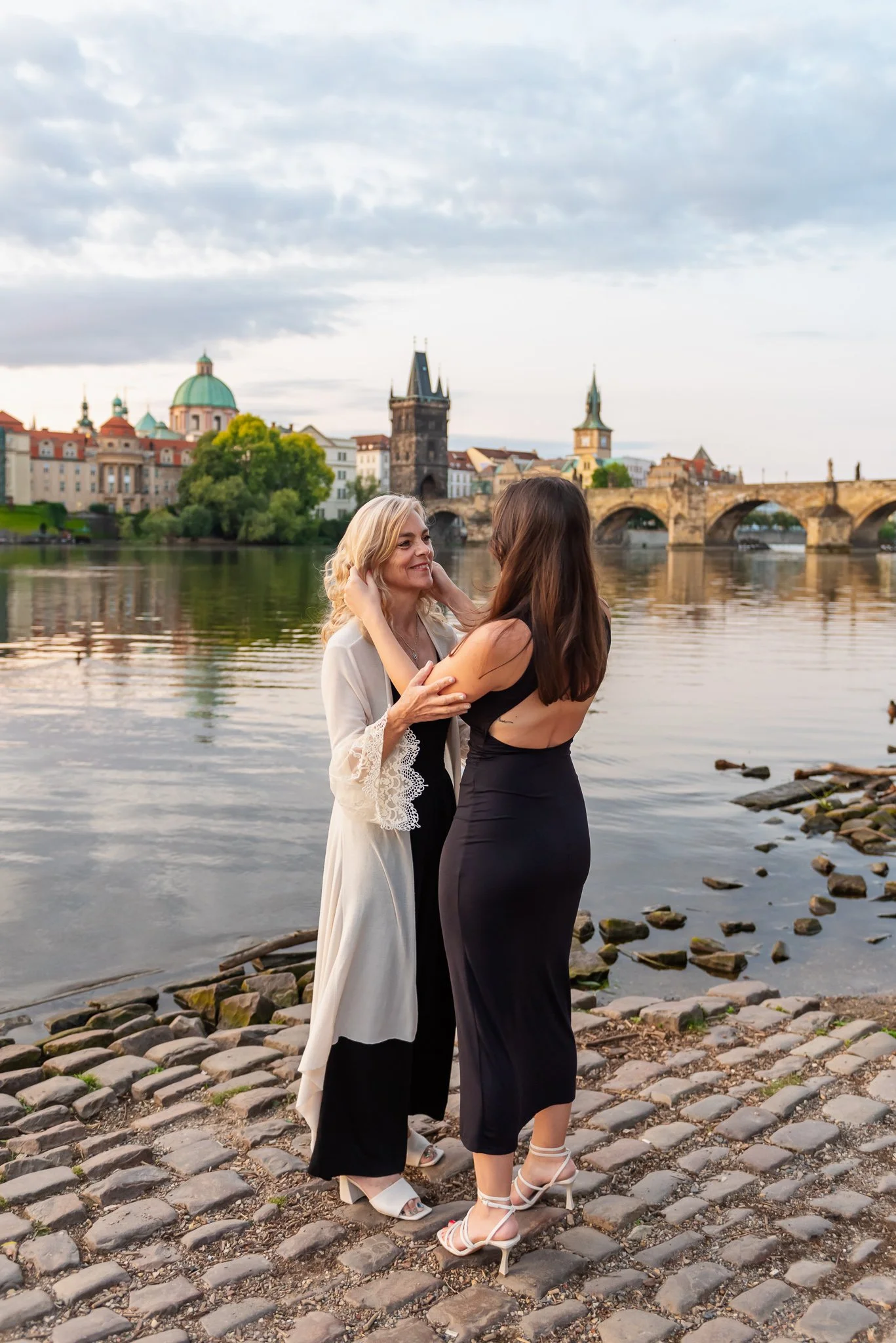 Two women, one with blonde hair wearing a white dress with lace sleeves and the other with brown hair wearing a black backless dress, standing close together on a cobblestone riverbank, smiling and touching each other's faces, with historic buildings and a bridge over a river in the background at dusk.