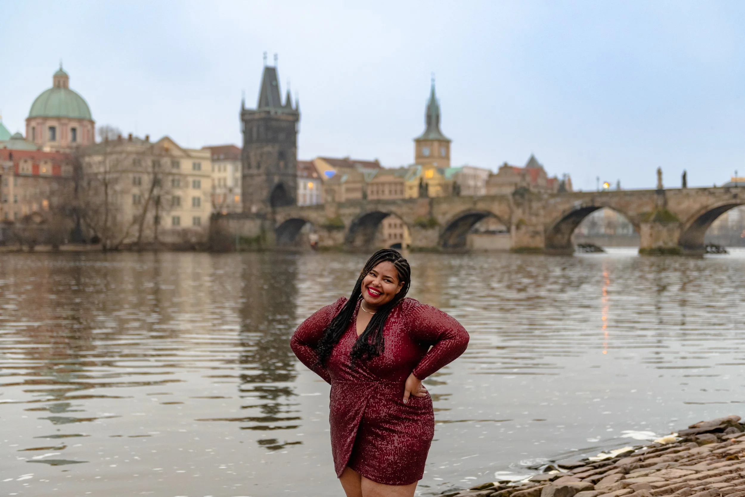 A woman in a sparkly red dress smiling by the river with historical European buildings and a stone bridge in the background.