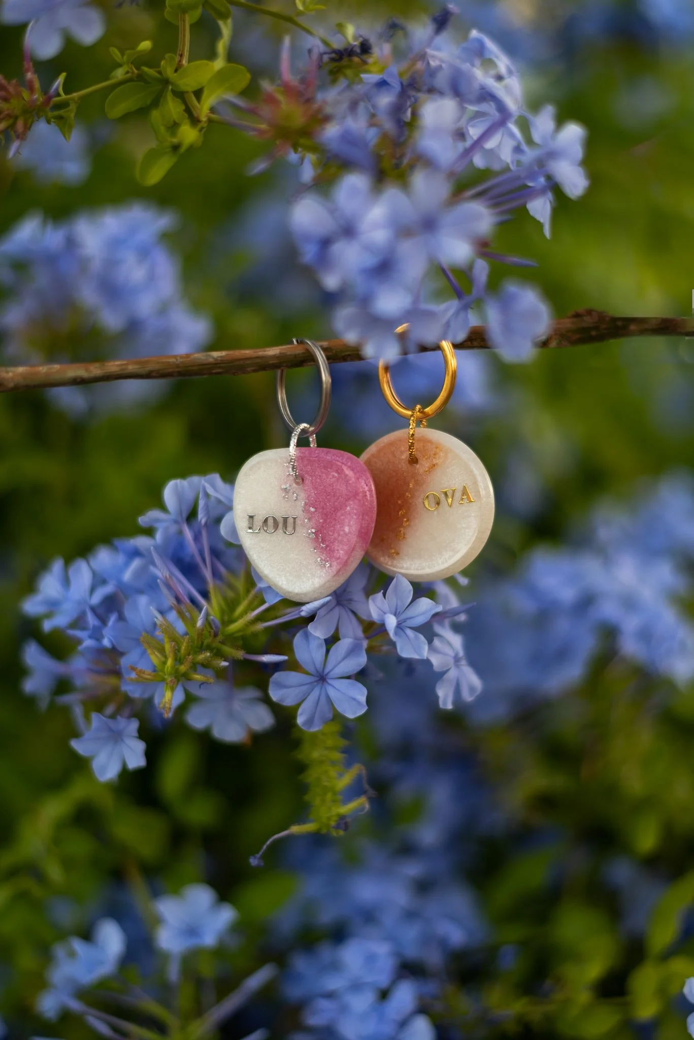 Two decorative keychains hanging on a thin branch among blue flowers with green leaves in the background. One keychain is shaped like a pink and white shell with the name 'LOU' engraved, and the other is round, beige, with the name 'OVA' engraved. Both keychains have small gold or silver rings.