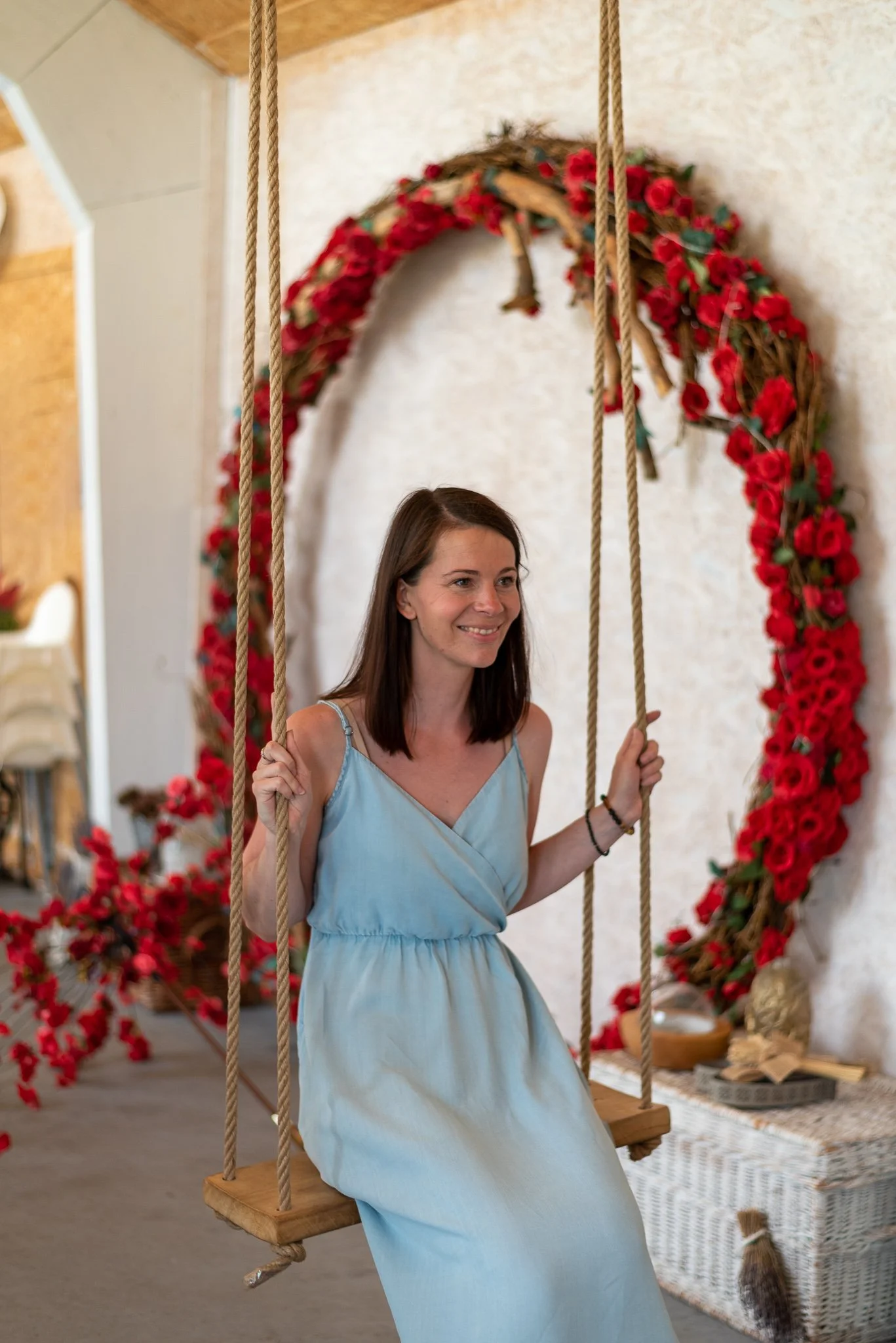 A woman smiling while sitting on a wooden swing with ropes, in front of a large floral wreath made of red flowers and green leaves, indoors with a beige wall and decorative items in the background. Personal branding photography in Prague.
