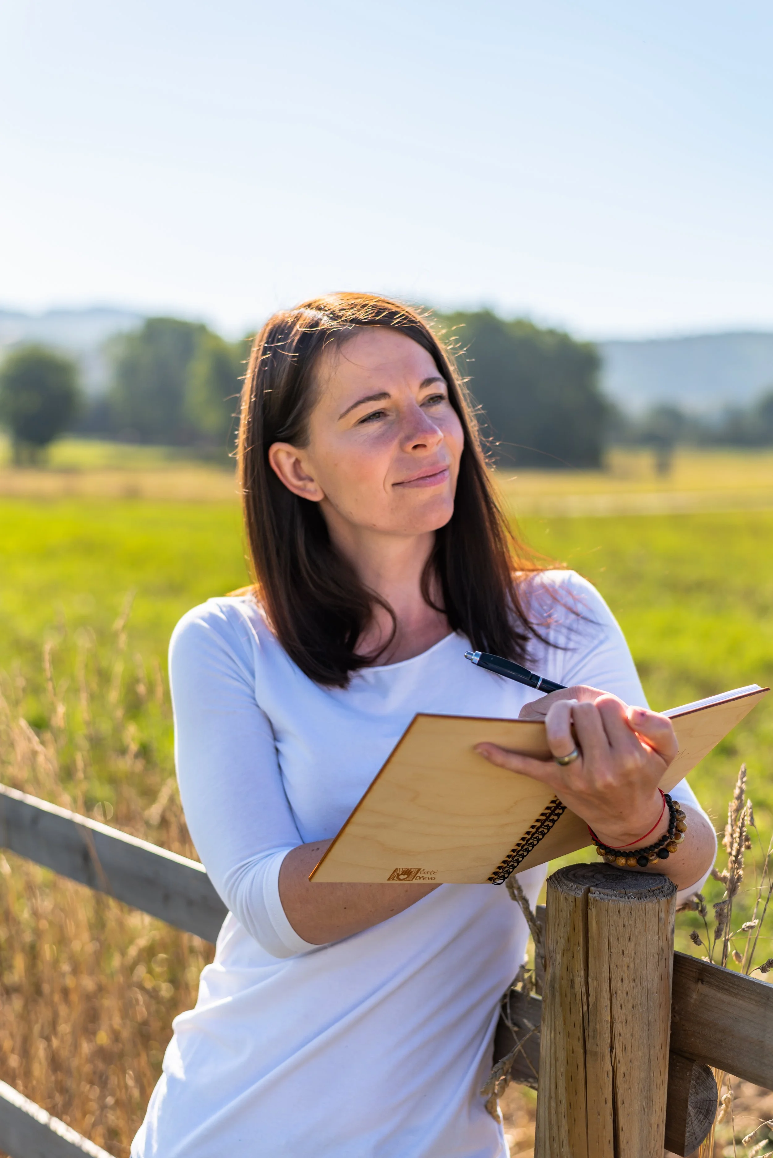 A woman with long brown hair, wearing a white shirt, is standing outdoors in a field with tall grass and trees in the background. She is holding a notebook and pen, and appears to be writing notes while looking into the distance with a slight smile. The weather is sunny and clear.