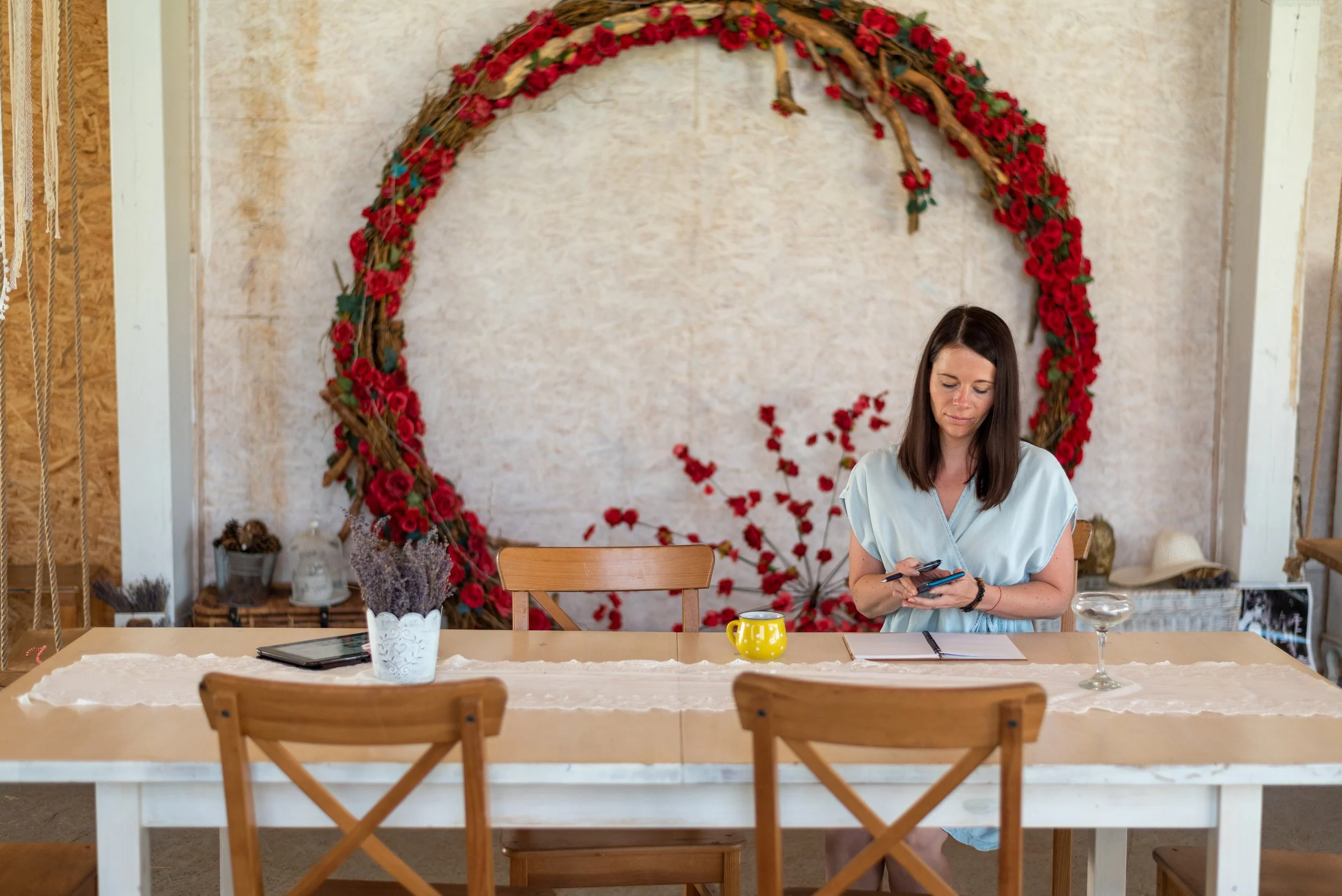 Woman sitting at a wooden table with a notepad, a yellow mug, and a glass, in front of a large floral wreath decor on the wall.