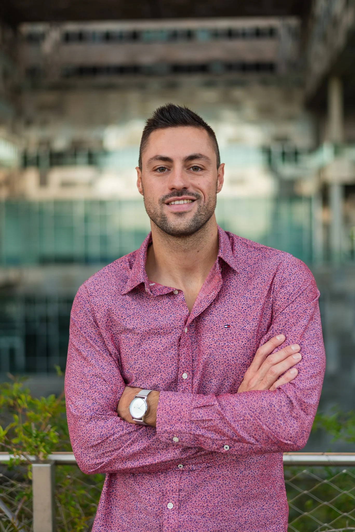 A man in a pink patterned shirt standing outdoors with arms crossed, smiling, with a modern building and water in the background.