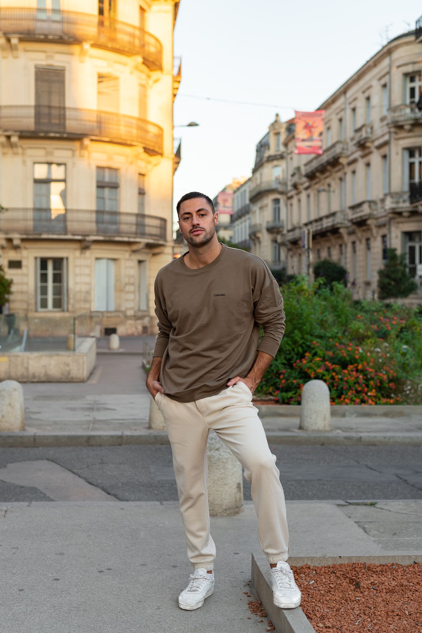 A young man stands with hands in his pockets on a city street at sunset, surrounded by European-style buildings and greenery.