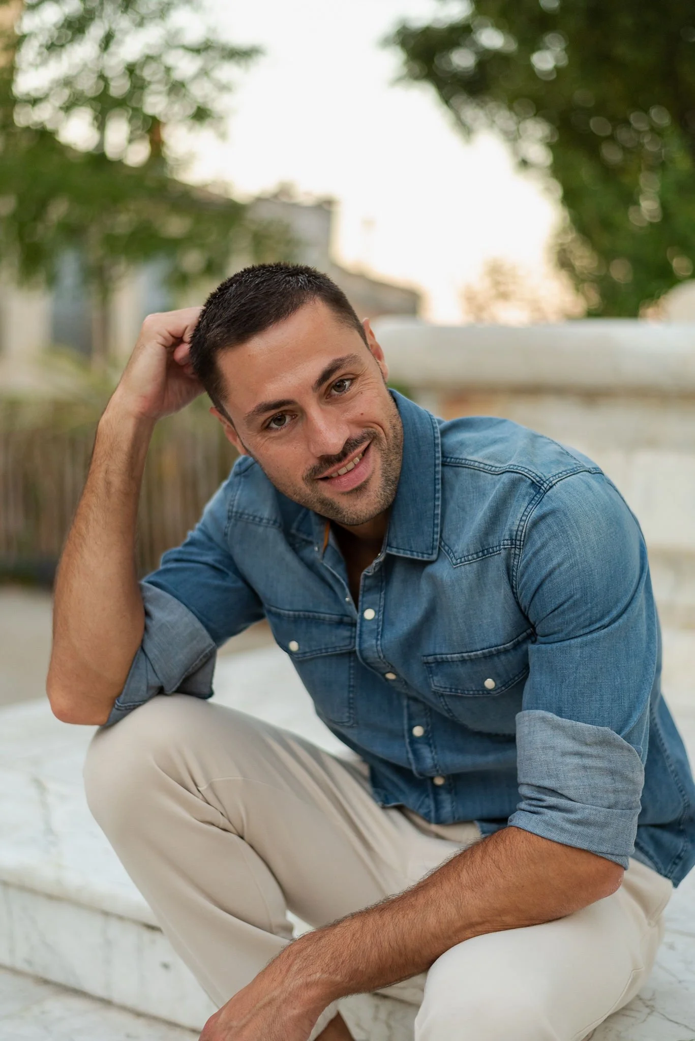 A man sitting outdoors on a white marble surface, smiling, with his left hand resting on his head. He is wearing a denim shirt and beige pants, with trees and a blurred background behind him.