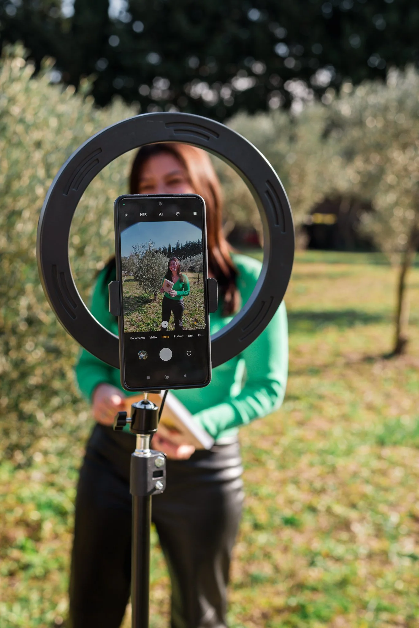 A woman in a green sweater holding a notebook, standing outdoors among trees and grass, as a smartphone on a tripod takes her picture through a ring light.