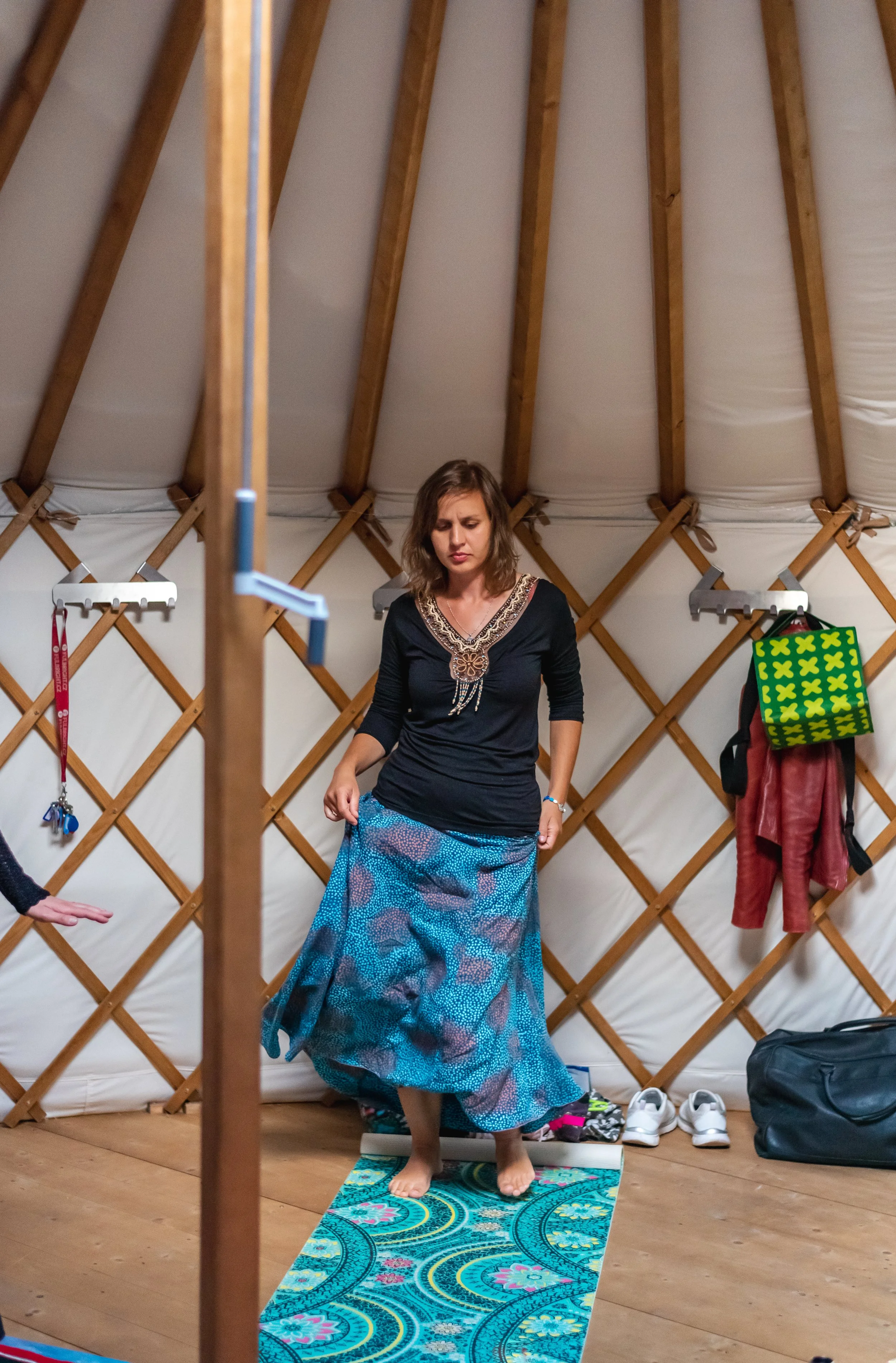 Woman standing on a colorful yoga mat inside a yurt, adjusting her long skirt, with shoes and bags in the background. Woman circle in Prague.