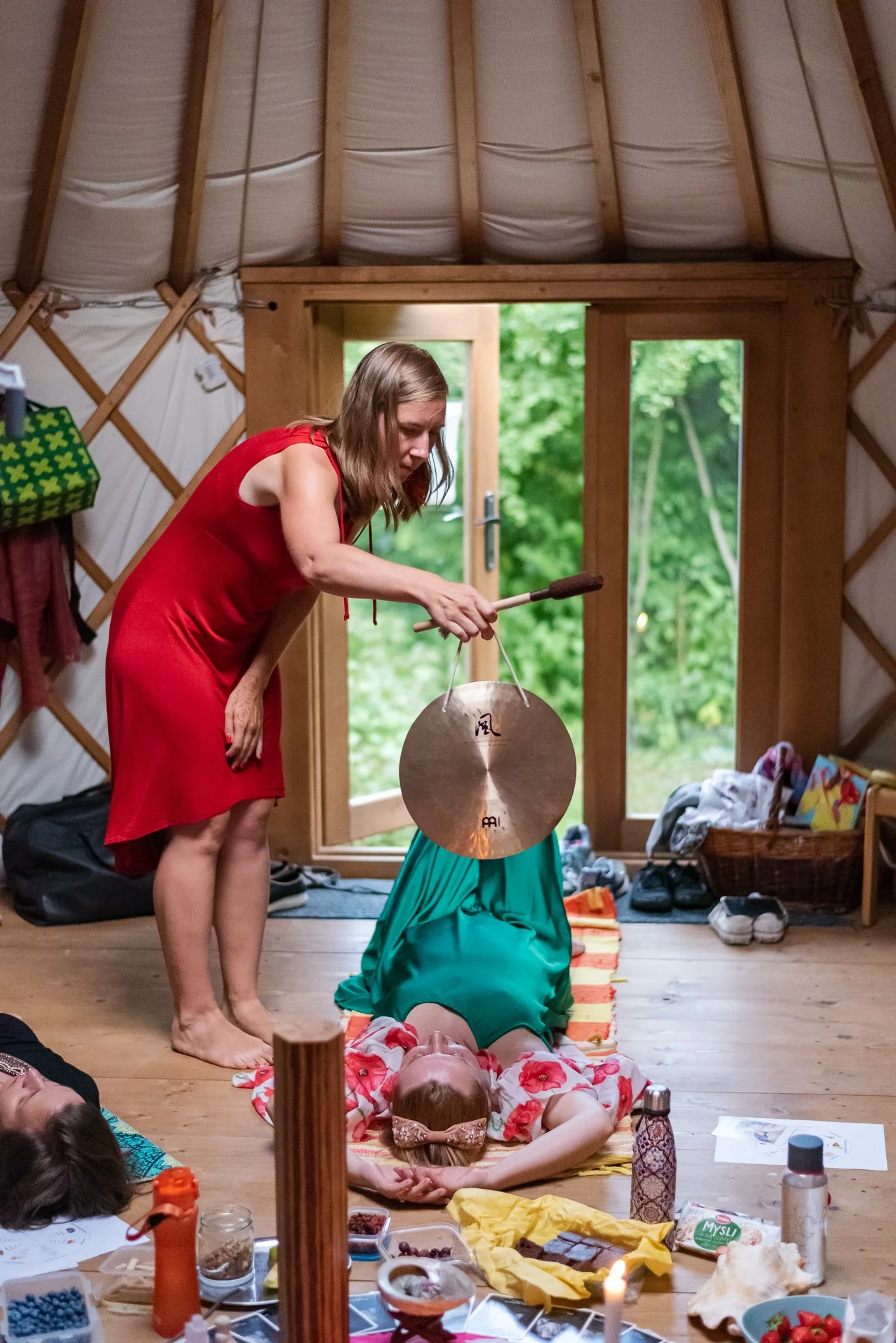 A woman in a red dress is playing a gong above a girl lying on the floor, inside a rustic wooden room with a door and windows showing greenery outside. Woman circle in Prague.