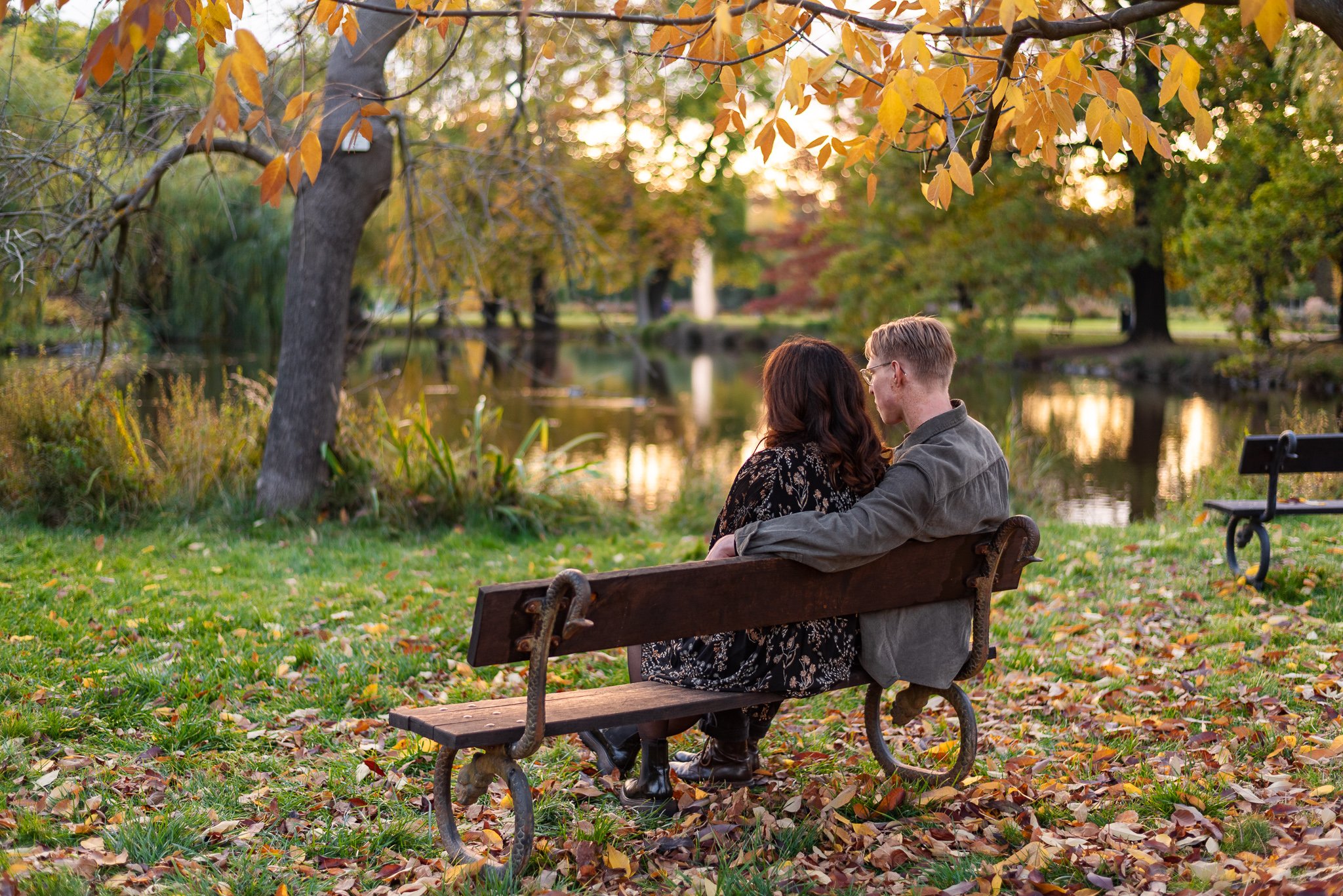 A couple sitting on a park bench near a pond during autumn in Stromovka park, surrounded by fallen leaves and colorful trees at sunset.