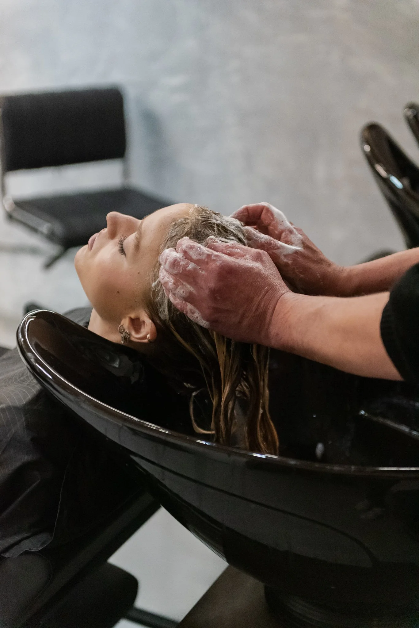 Person receiving a head massage or shampoo at a salon, with their head leaning back in a black salon sink and someone washing or rinsing their hair. Content creation for hairdresser in Prague.