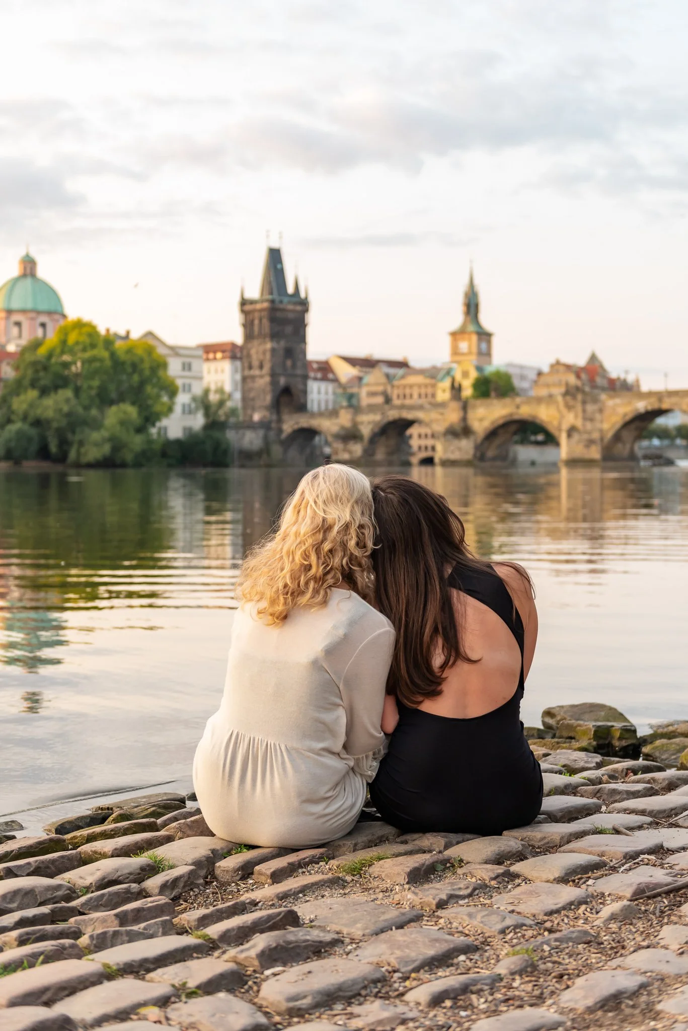 Two women sitting on cobblestone riverside at sunset, with historic Charles bridge and cityscape in the background.
