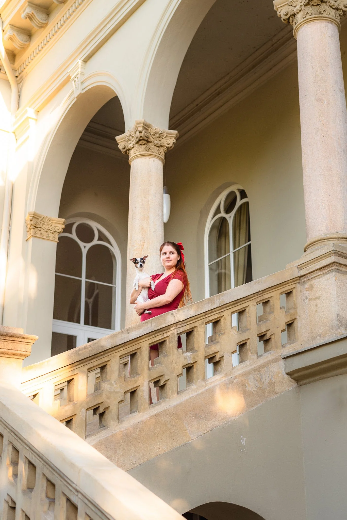 A woman with red hair and a red dress holding a small dog, standing on a stone balcony of a classical-style building with arched windows and ornate columns.