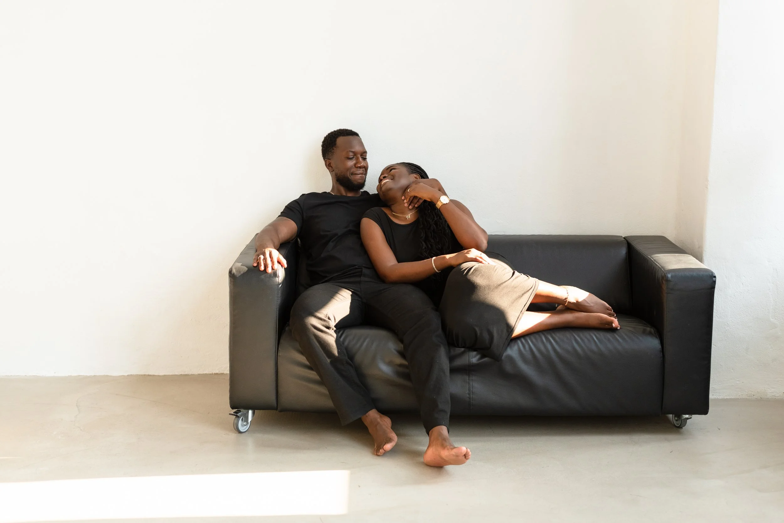 A happy couple sitting on a black leather couch, looking at each other and smiling, in a minimalist room with plain white walls.