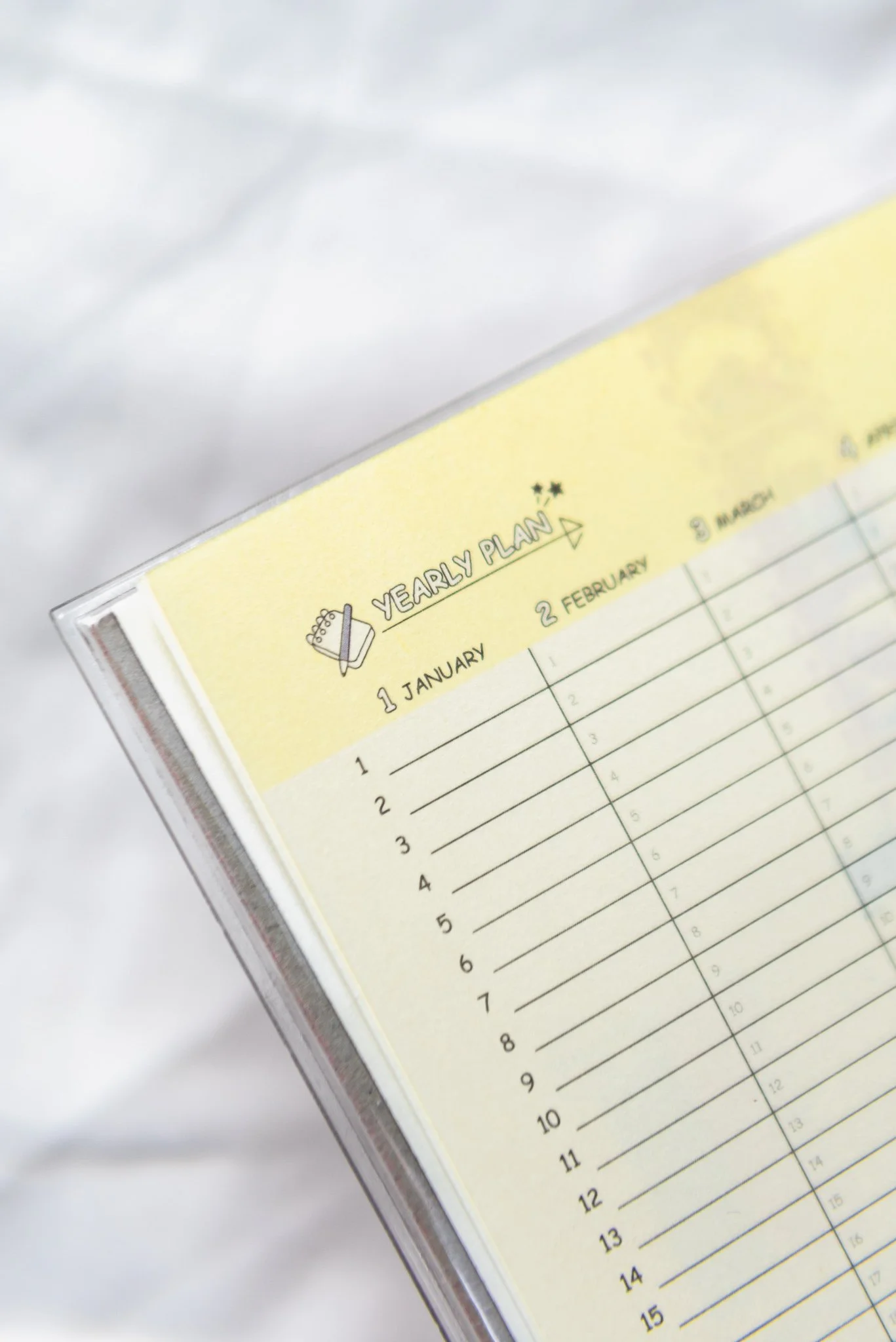 Close-up of a yellow yearly planner page with handwritten dates and lines for planning, partially inside a clear plastic cover, on a white background.