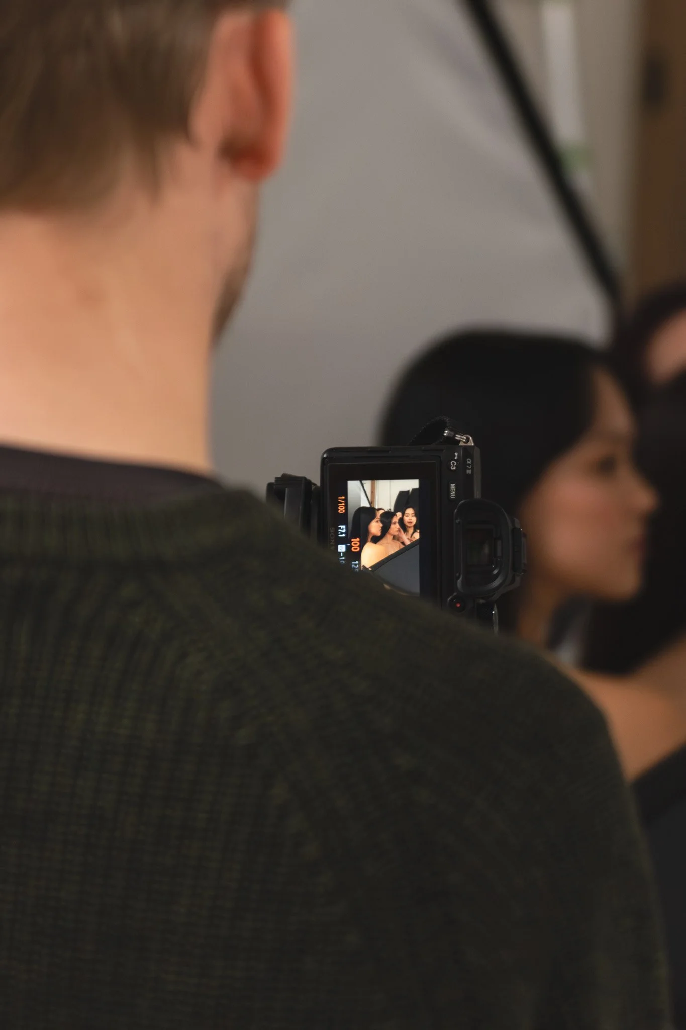 A person with short hair, seen from behind, taking a photo or recording with a digital camera. The camera's screen shows a woman with dark hair and light skin, posing against a dark background.