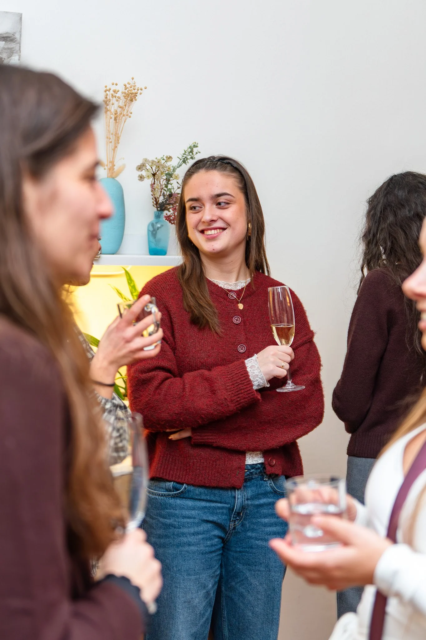 A group of women socializing at a gathering, one woman in the center smiling, holding a glass of champagne, wearing a red sweater and jeans, with others holding drinks around her, indoor setting with white wall and decorative vases with dried plants.