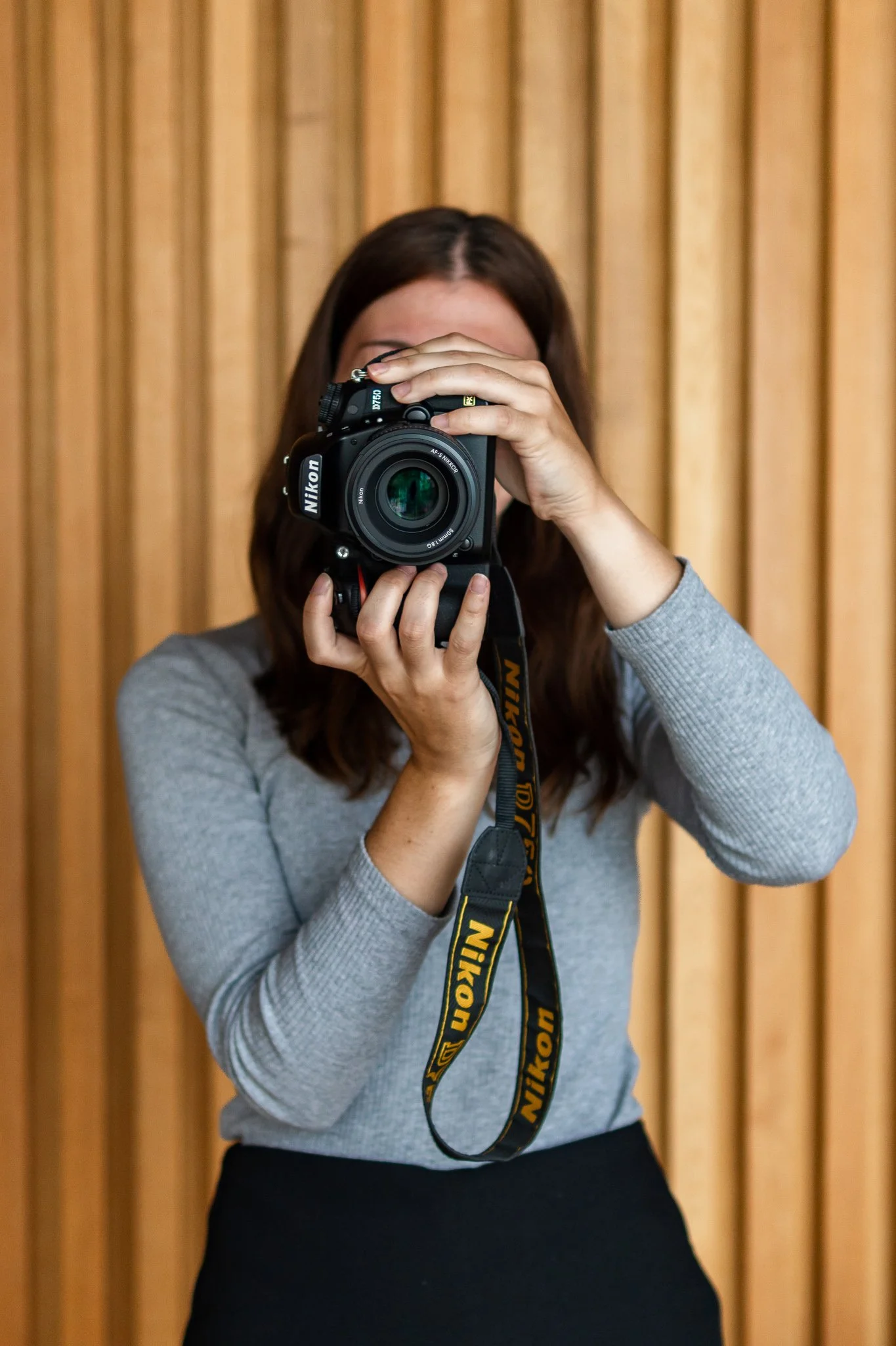 A woman with brown hair in a gray long sleeve shirt holds a Nikon DSLR camera and takes a photo indoors in front of a wooden slat wall.