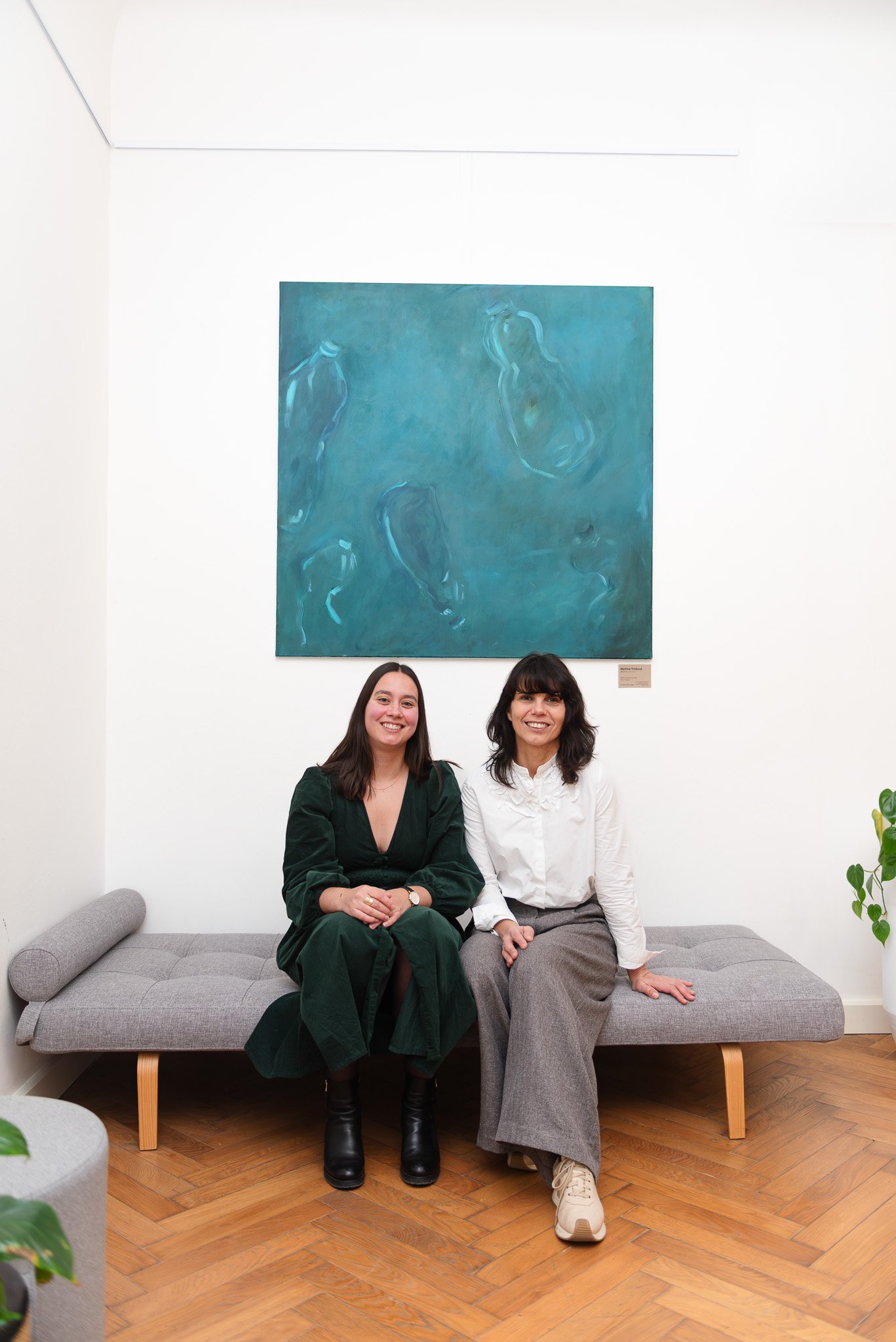 Two women sitting on a gray sofa in an art gallery, smiling at the camera, with a blue abstract painting on the white wall behind them, and a small plant on the right side of the image.