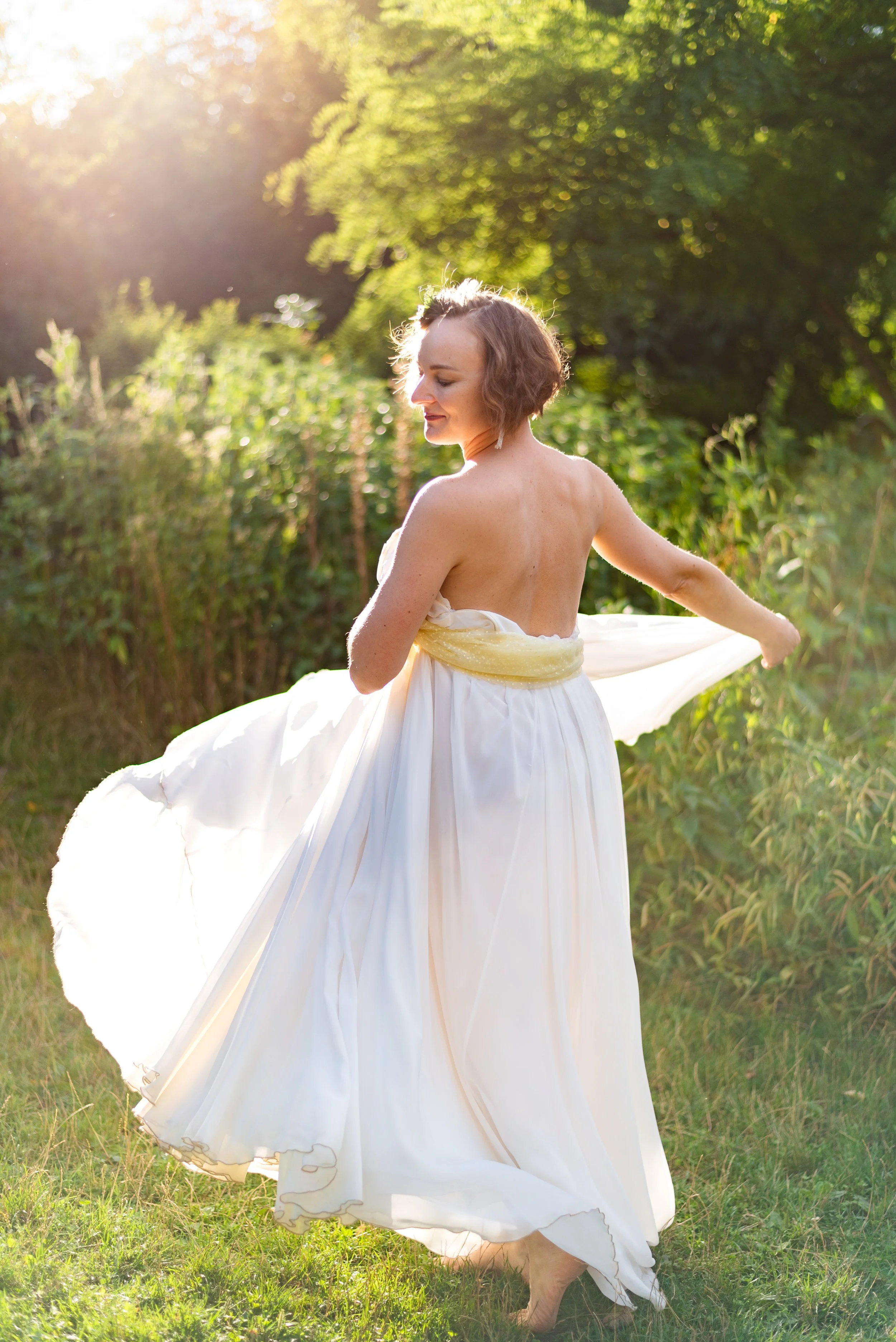 A woman in a flowing white dress with a yellow sash dances barefoot in a sunlit outdoor setting surrounded by greenery.