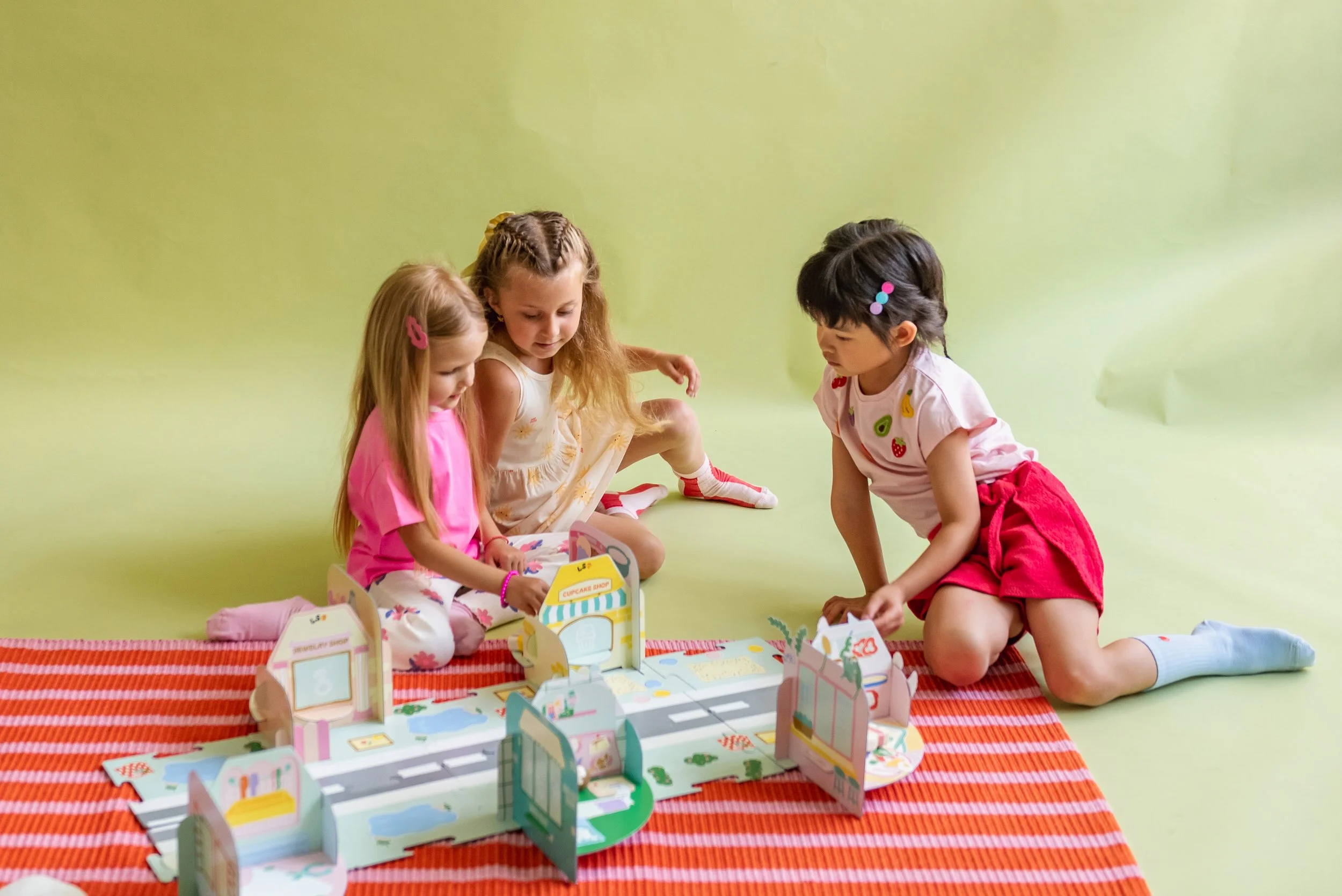 Three young girls playing with a colorful cardboard play set on a striped orange rug, with a plain green background.