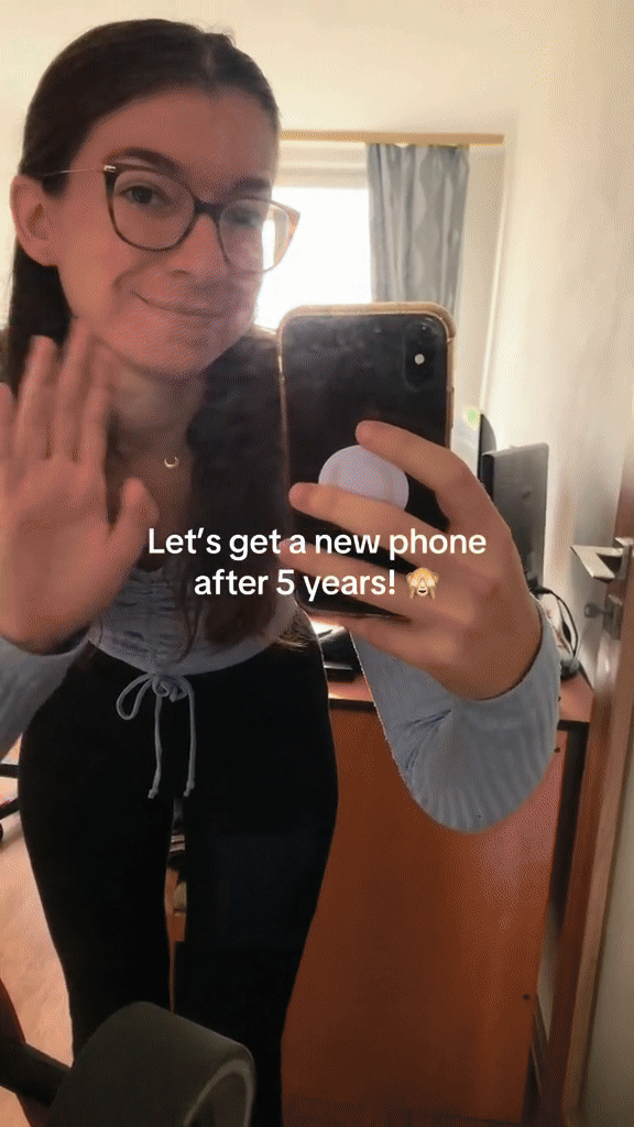 Young woman in glasses smiling and waving, taking a selfie in a mirror, with text overlay that reads "Let's get a new phone after 5 years!"