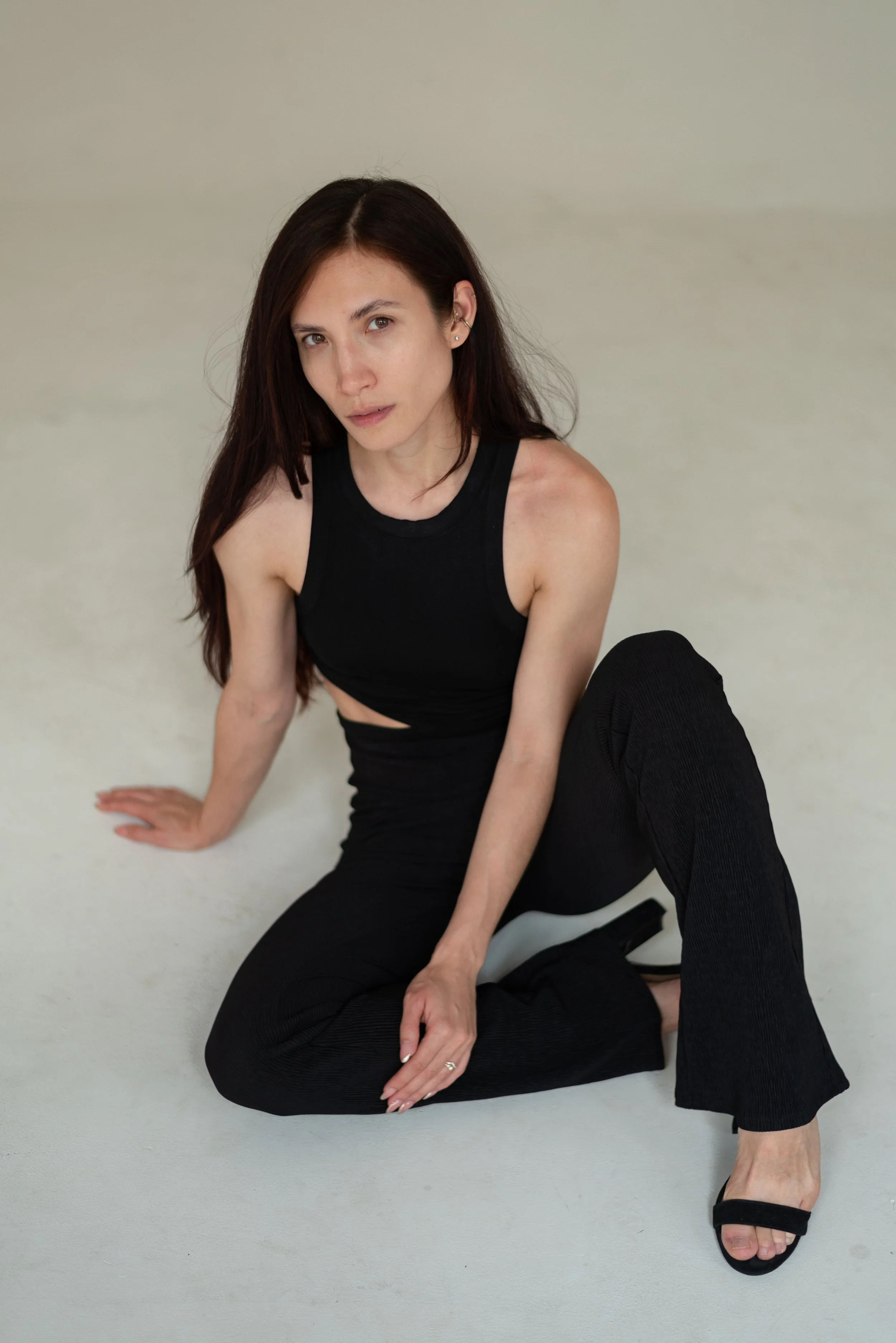 A woman with long brown hair sitting on a light-colored floor, wearing a black sleeveless top, black pants, and black high-heeled sandals, looking at the camera.