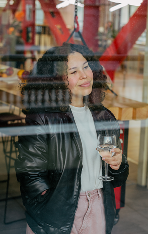 Young woman with curly hair holding a glass of white wine, looking out a window in a casual indoor setting.